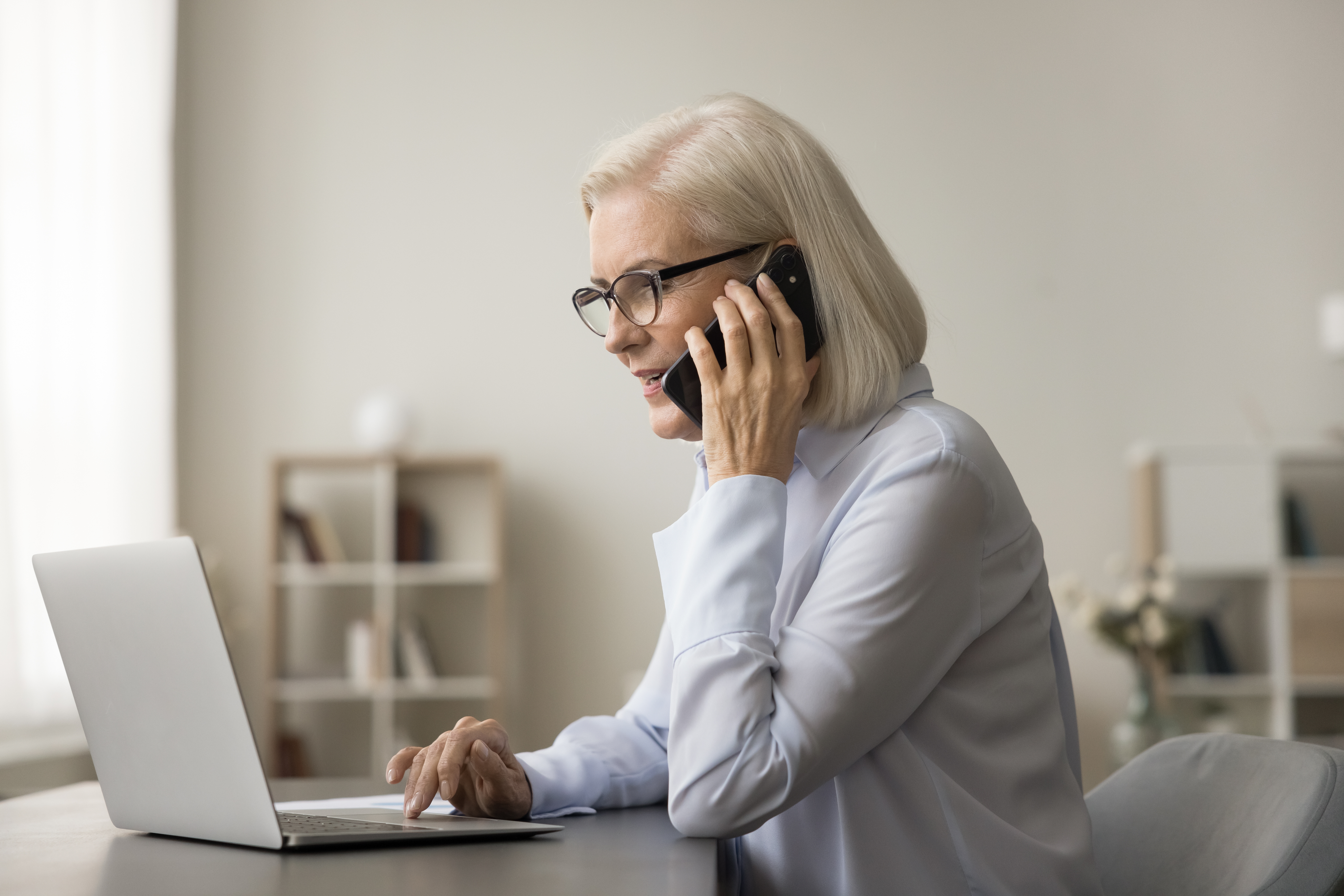 Mujer mayor hablando por teléfono frente a un portátil | Foto: Shutterstock