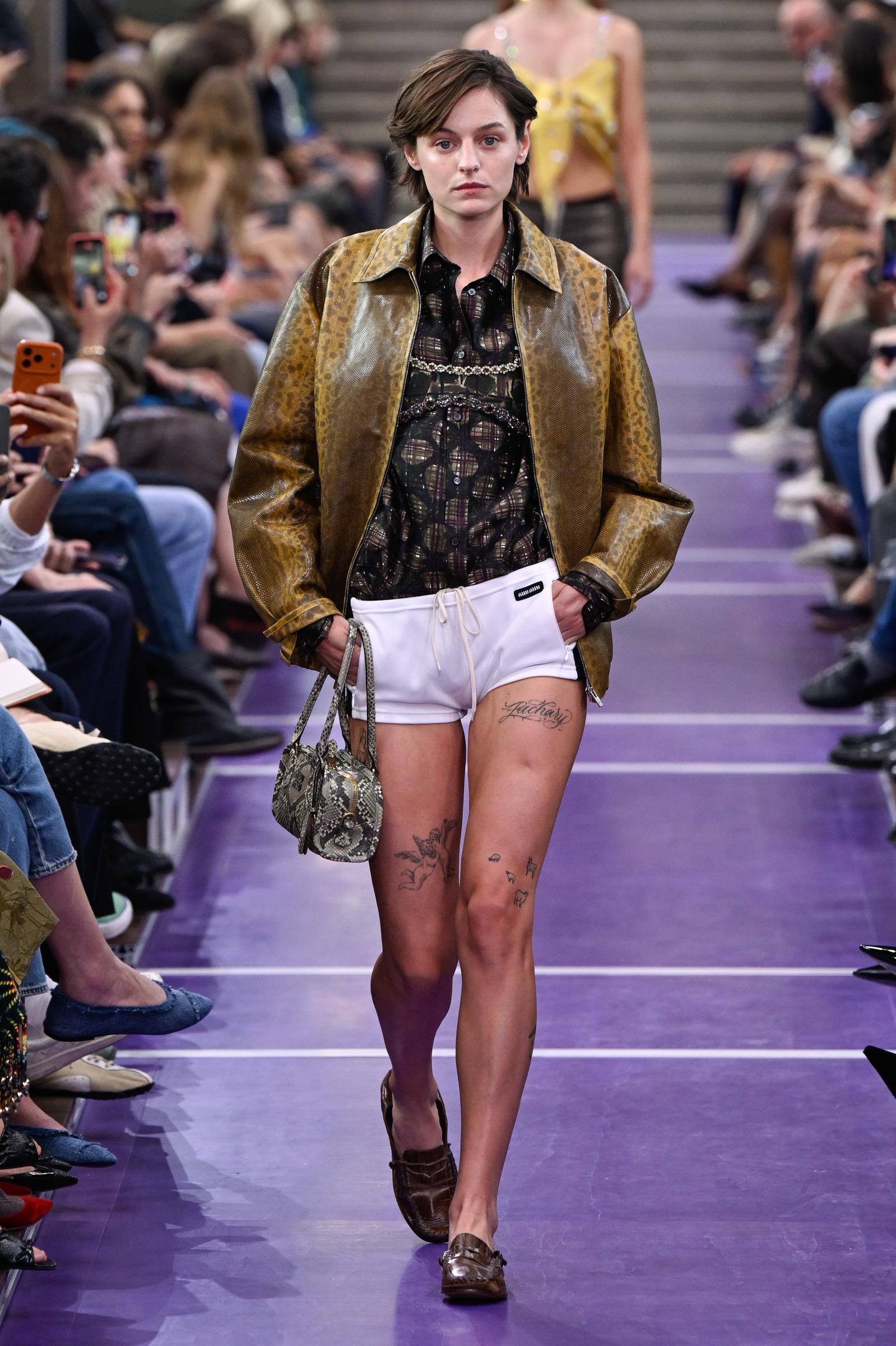 Emma Corrin desfila por la pasarela de Miu Miu con una chaqueta de cuero marrón con textura sobre un top negro transparente, combinada con unos pantalones cortos blancos con cordón, un bolso estampado y unos mocasines marrones. | Fuente: Getty Images