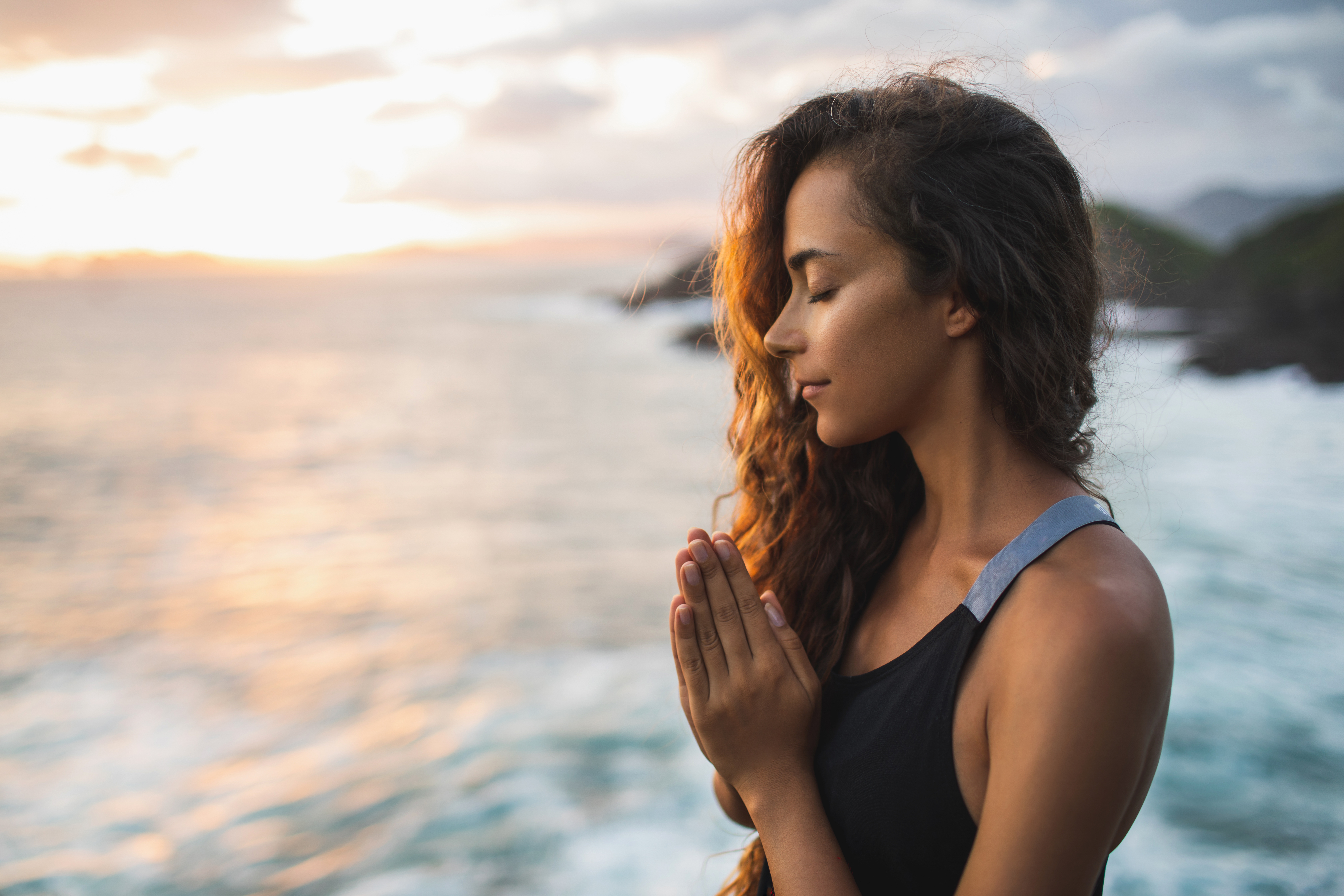 Una mujer de pie cerca del océano con las manos en posición de oración con los ojos cerrados. | Foto: Shutterstock