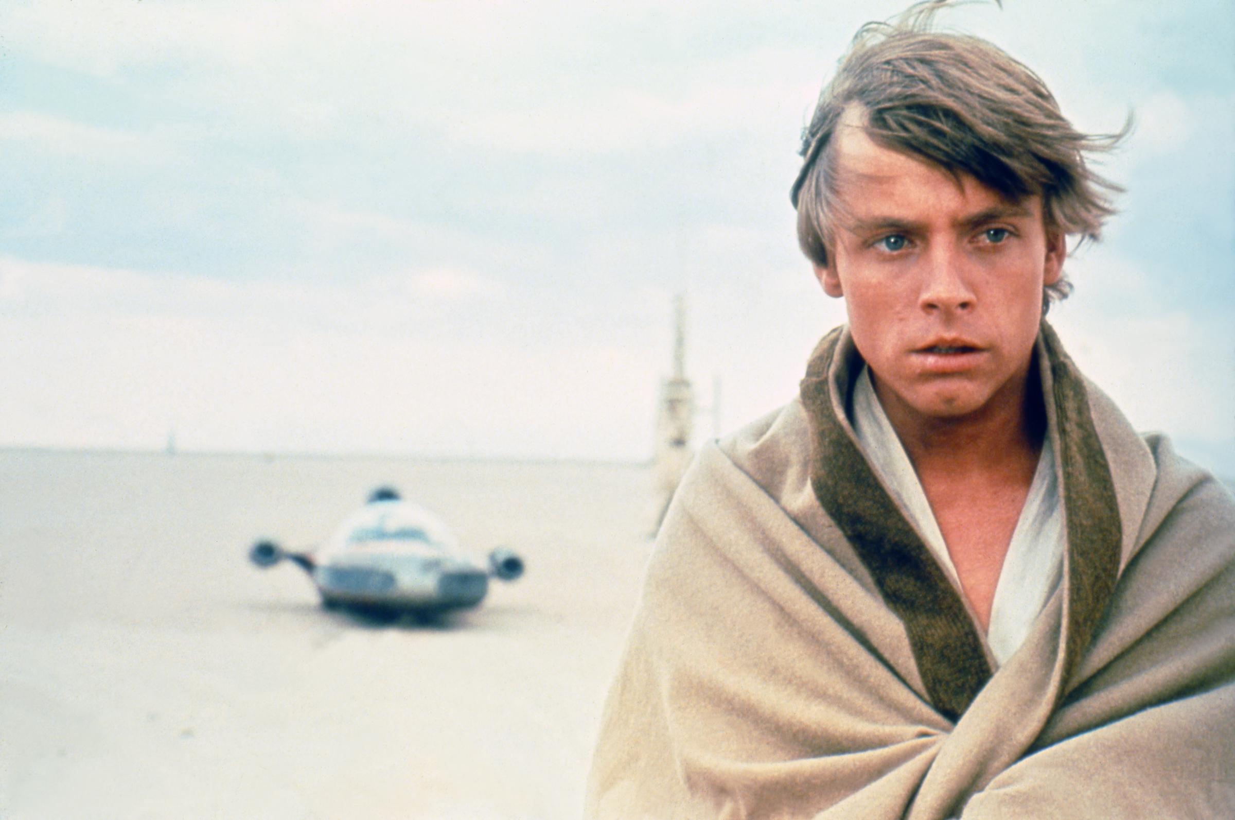 El actor se mantuvo de pie ante el árido paisaje desértico durante la producción de "Star Wars: A New Hope", con el landspeeder del personaje visible a lo lejos. Envuelto en prendas de color claro bajo el cielo abierto, personificó al joven héroe antes de que el público supiera su nombre. La foto de producción ofreció otro vistazo claro de su apariencia antes del accidente de enero de 1977 que derivó en un tratamiento reconstructivo para sus lesiones faciales. | Fuente: Getty Images