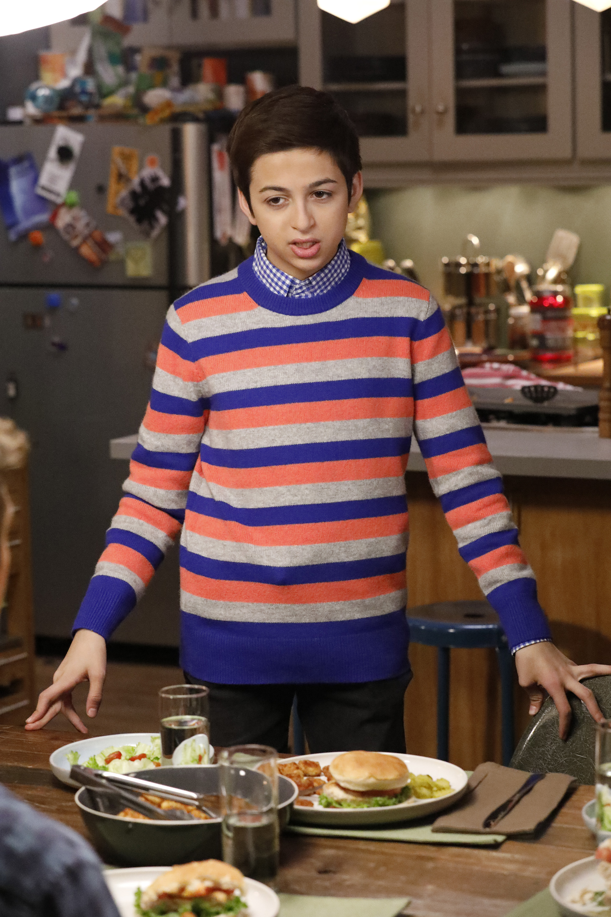 J.J. Totah interpreta a Michael en el episodio de Champions "Vincemas", usando un suéter a rayas de colores sobre una camisa a cuadros azul mientras está de pie junto a una mesa de comedor en una cocina. | Fuente: Getty Images