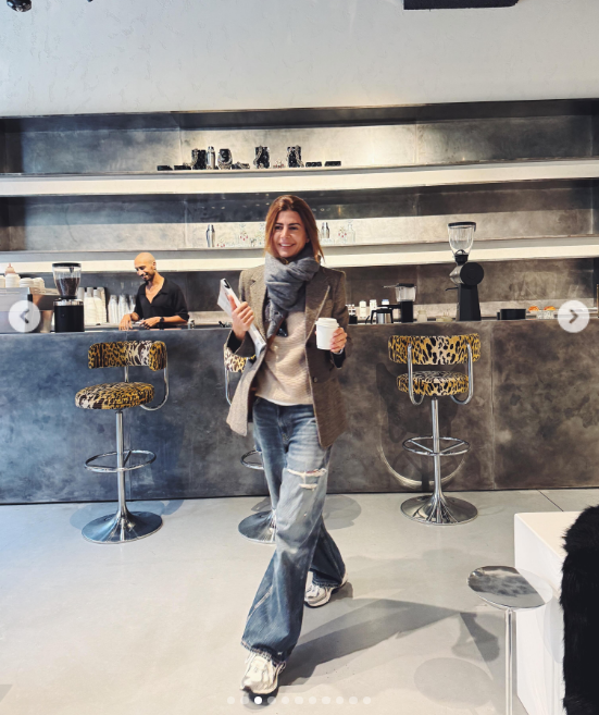 Juliana Awada camina sonriente por una moderna cafetería llevando un café para llevar y una revista bajo el brazo, vestida con blazer marrón, bufanda gris envolvente, jeans anchos desgastados y zapatillas deportivas. | Fuente: Instagram/juliana.awada