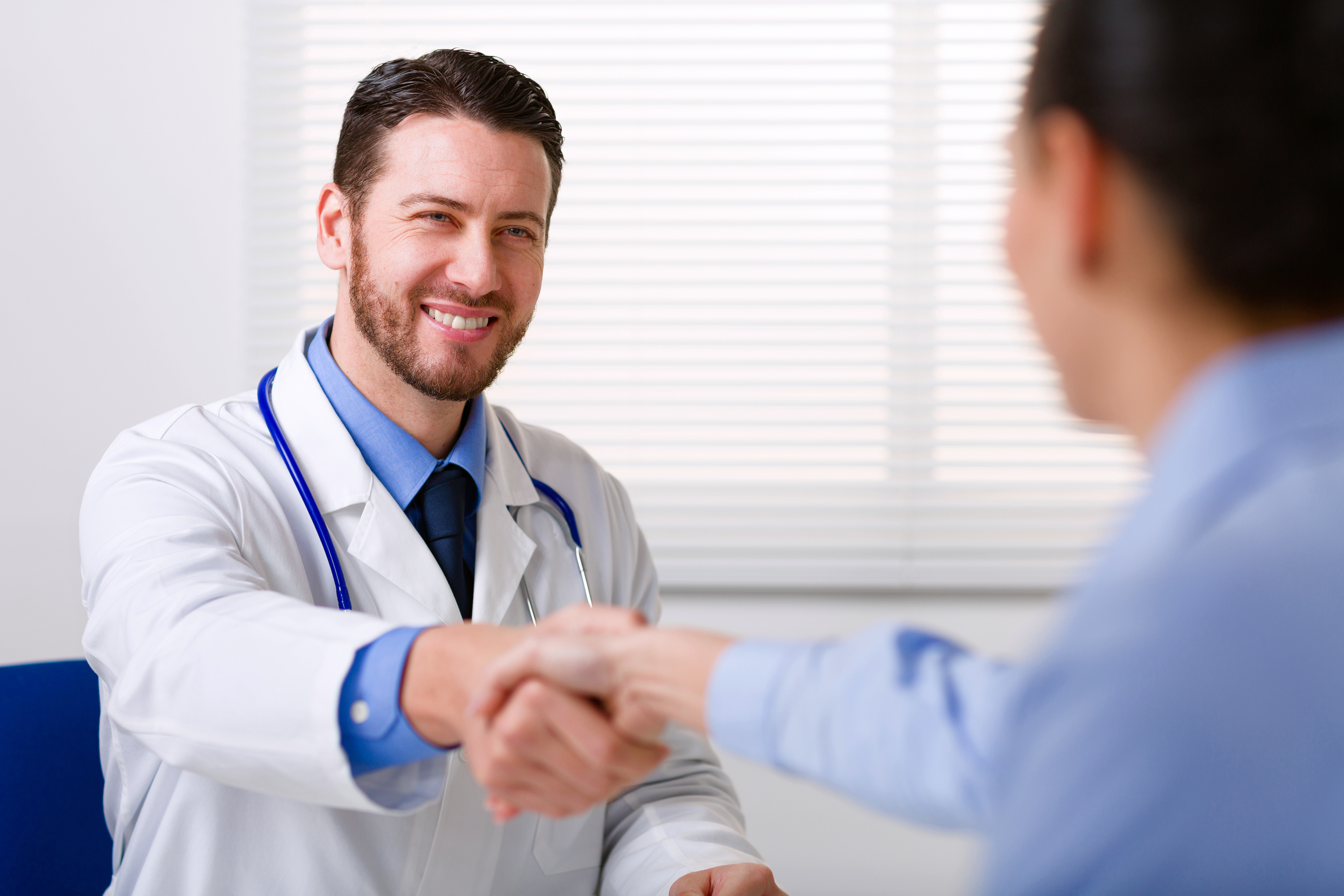 Médico con bata blanca sonriendo mientras estrecha la mano a una mujer. | Fuente: Shutterstock