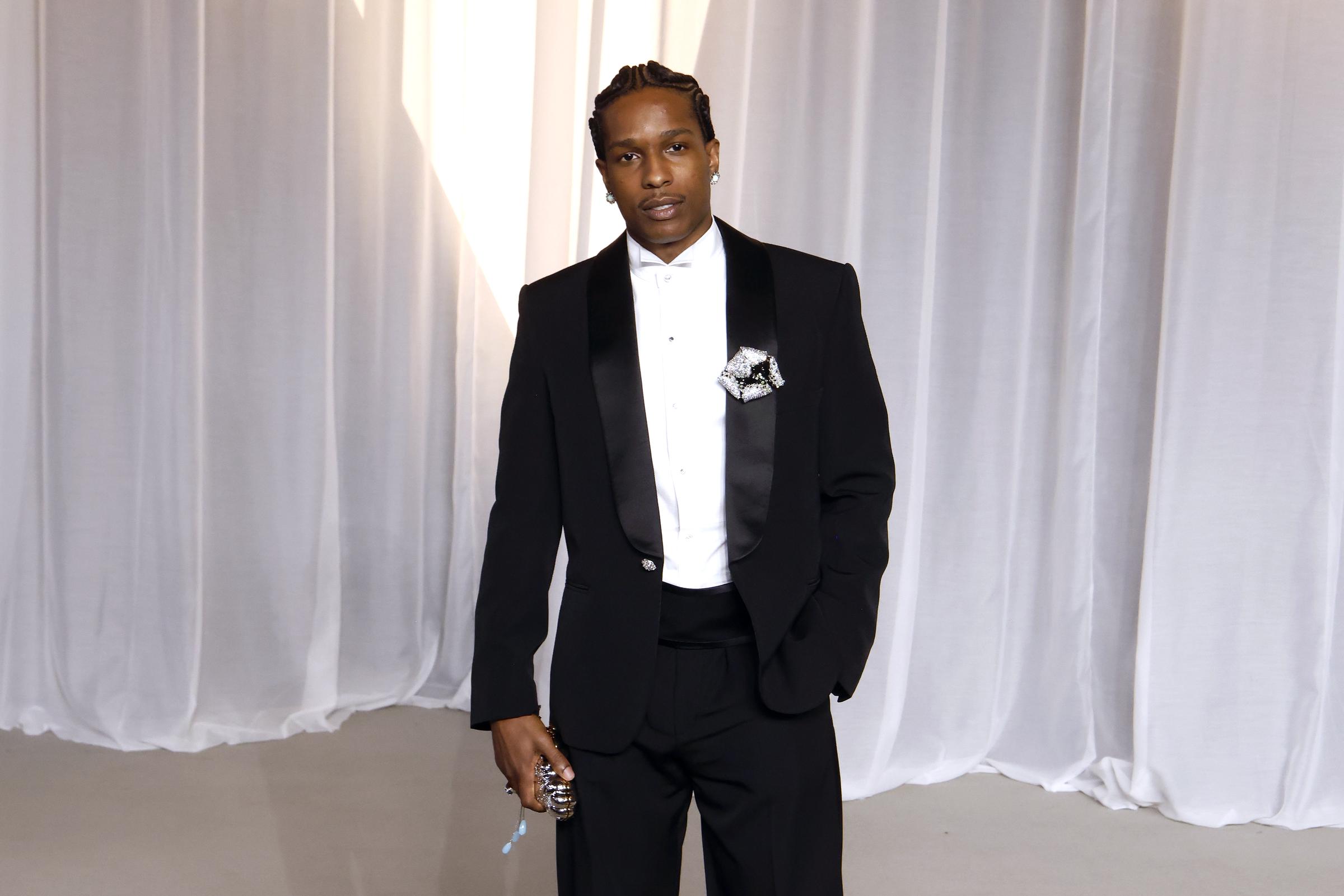 A$AP Rocky luce un look elegante y moderno de etiqueta con un esmoquin de líneas depuradas, confeccionado a medida con amplias solapas de satén tipo chal y una faja alta a la cintura. En lugar del corbatín tradicional, el rapero opta por una camisa blanca de vestir minimalista con cuello de alas y realza este conjunto clásico con un broche floral muy elaborado en la solapa, unas trenzas africanas muy cuidadas y un llamativo bolso de mano de noche.