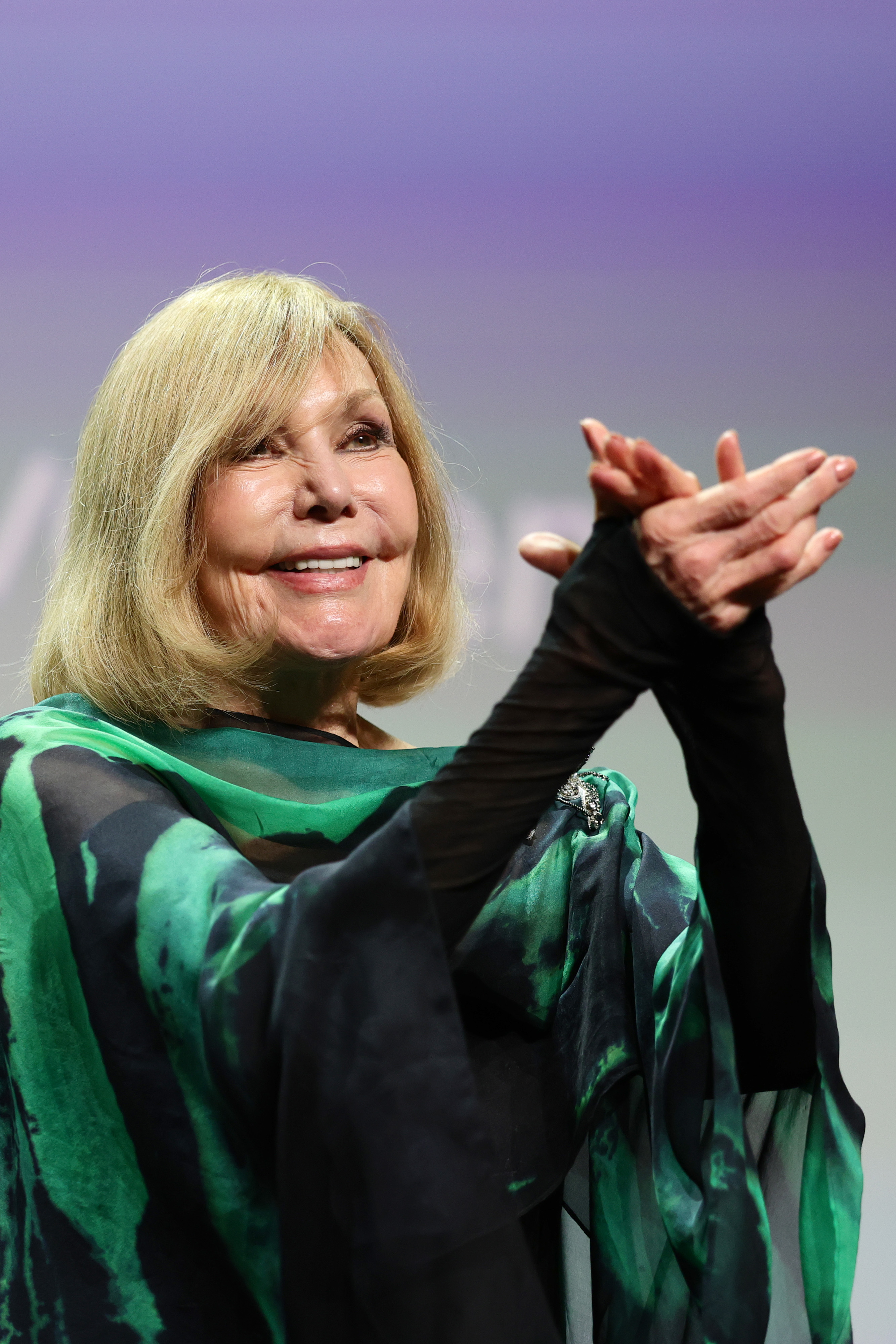 Kim Novak sonríe y aplaude durante la ceremonia de entrega del León de Oro a toda una carrera en el 82.º Festival Internacional de Cine de Venecia, el 1 de septiembre de 2025. Lleva un chal vaporoso en tonos verde azulado y verde sobre un conjunto negro mientras saluda al público. | Fuente: Getty Images