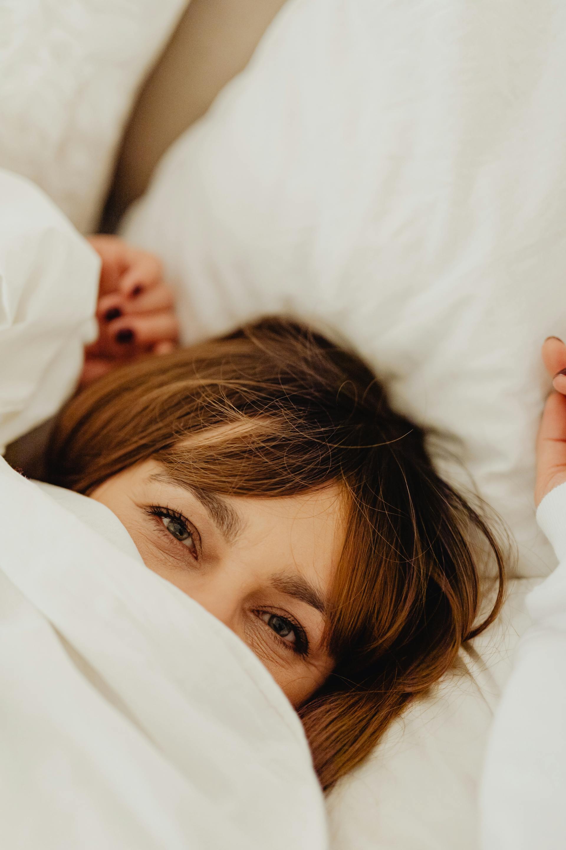 Una mujer tumbada en la cama con la cara oculta bajo una manta blanca | Fuente: Pexels