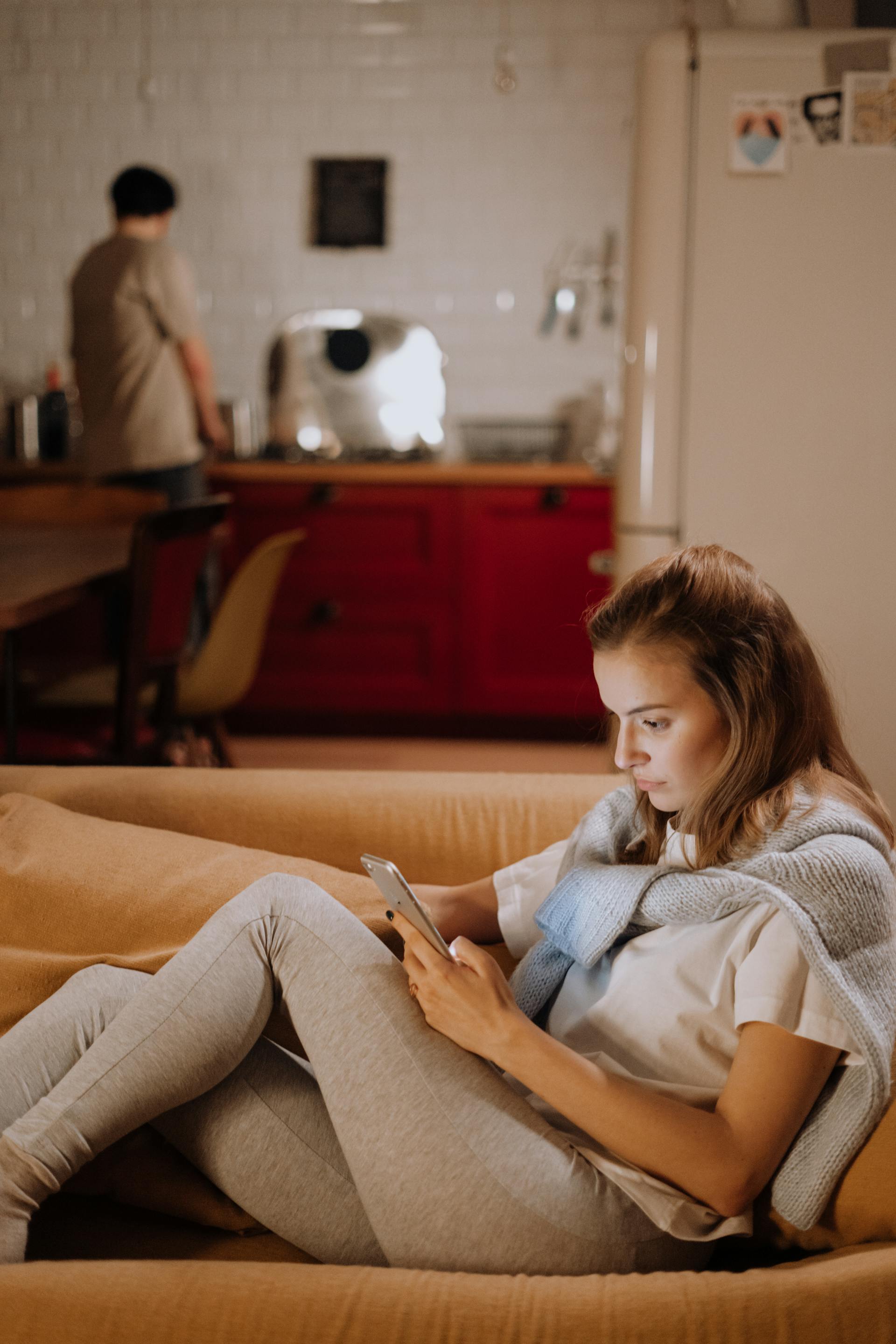 Una mujer sentada en el sofá y utilizando su teléfono mientras un hombre está de pie en la cocina | Fuente: Pexels