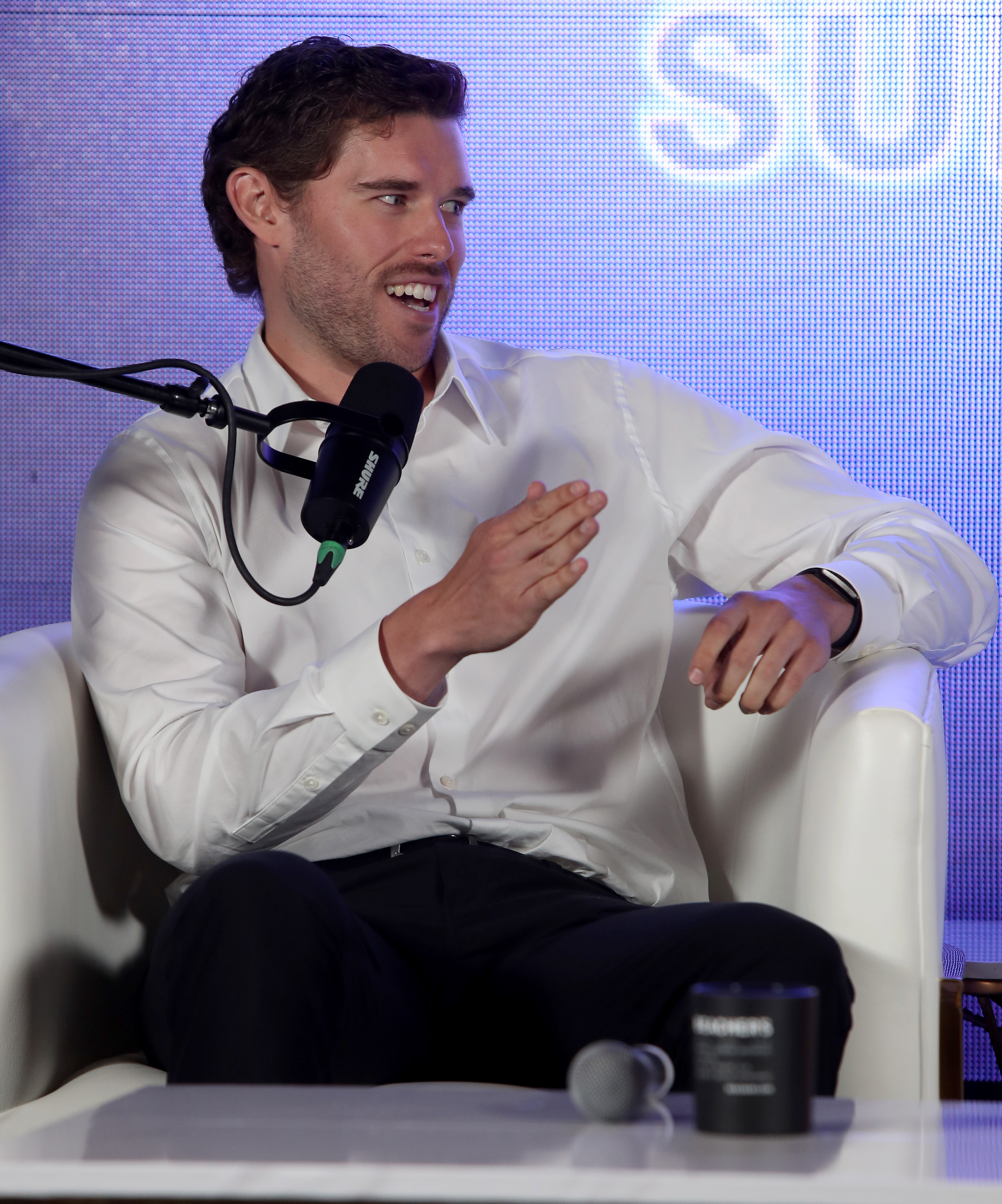 Christopher Schwarzenegger en plena discusión durante un panel en la Cumbre Beacher Vitality Happy & Healthy. Con una camisa blanca entallada y unos pantalones negros ajustados, su esbelta figura refleja los resultados de sus años de transformación sanitaria.