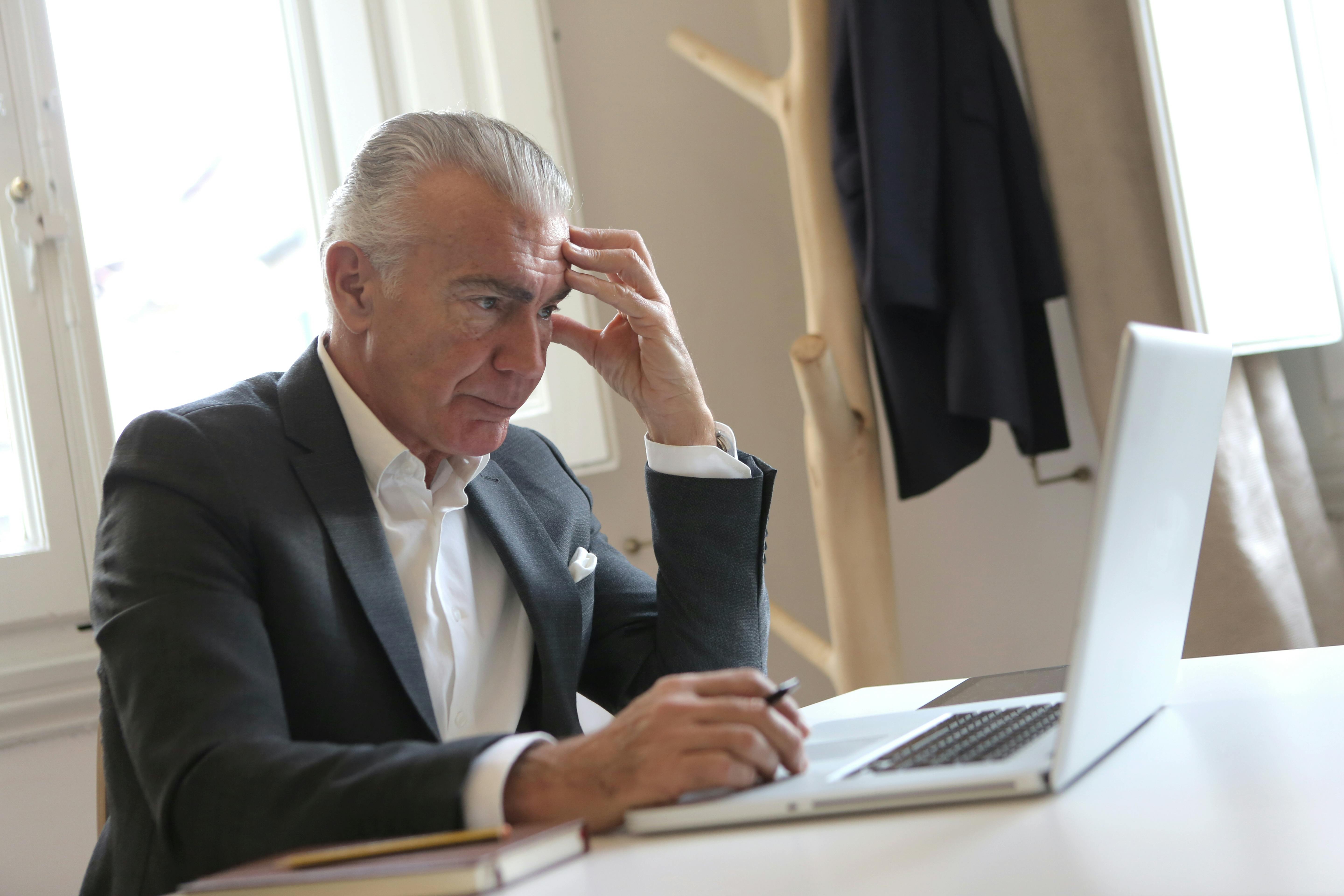 Un hombre mayor estresado sentado con la mano en la sien mientras sujeta un bolígrafo y mira su portátil | Foto: Pexels