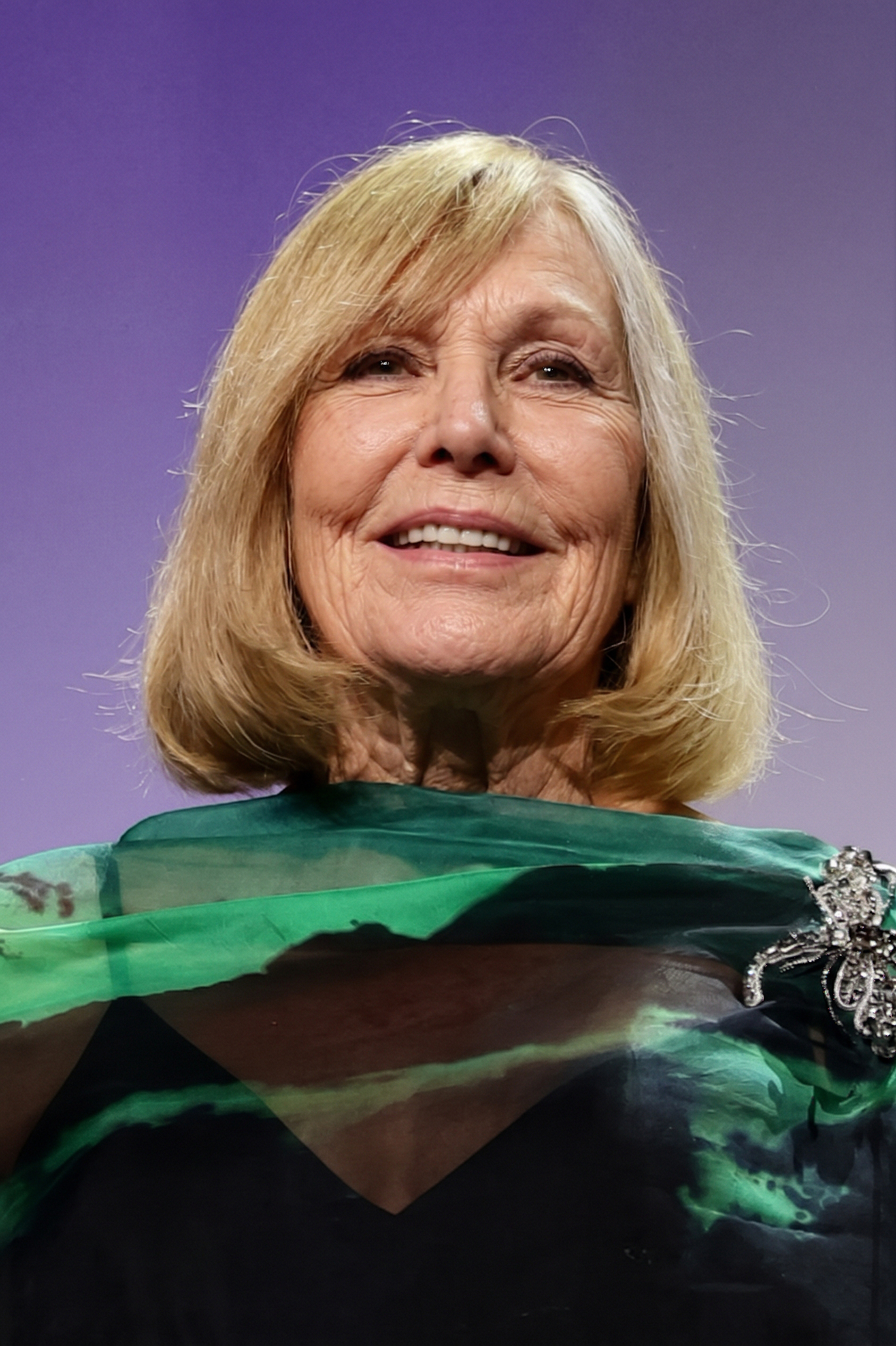Kim Novak sonríe con calidez con un conjunto verde y negro de tela transparente y un broche con pedrería en el hombro, sobre un fondo morado suave. | Fuente: Gemini