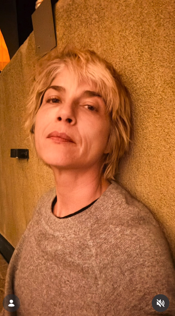 Selma Blair mira hacia arriba en un retrato relajado tomado desde un ángulo bajo, con la cabeza apoyada contra una pared de tierra con textura, el 11 de octubre de 2026. La cálida iluminación ambiental del interior le da un tono suave a su corte de pelo rubio en capas y a su jersey de cuello redondo en gris jaspeado. | Fuente: Instagram/selmablair