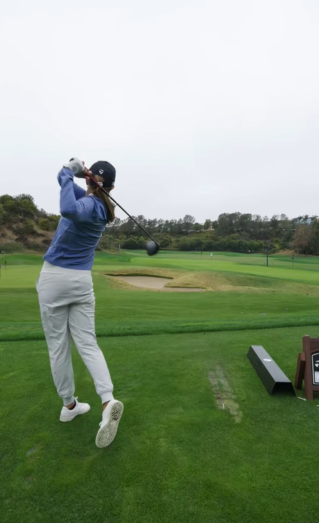 Kai Trump termina su swing en el campo de golf, vestida con un top azul, pantalones blancos y gorra negra en un día nublado | Fuente: Tiktok/thekaitrumpgolfer