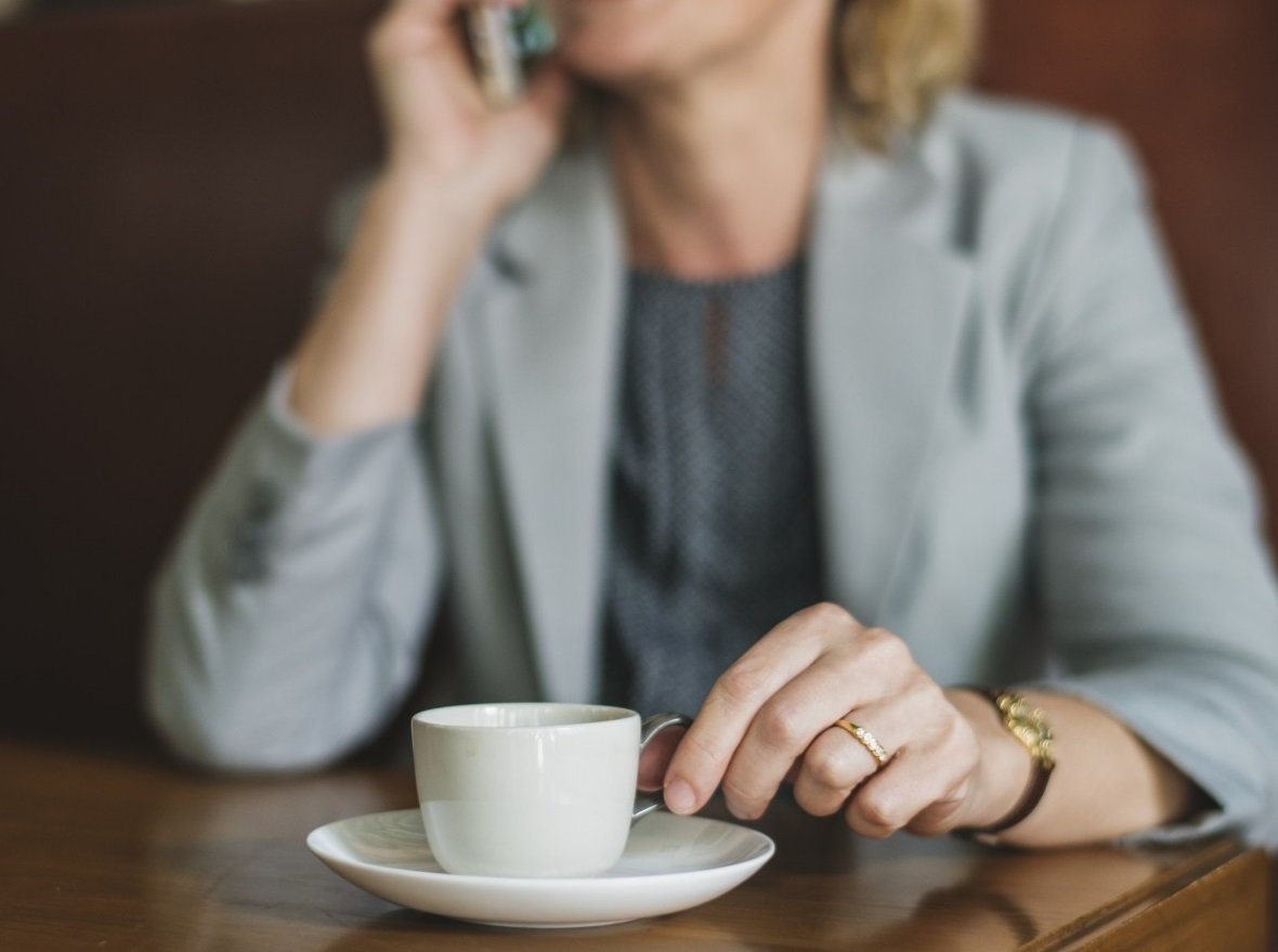 Una mujer hablando a través de un teléfono celular sentada frente a una mesa con una tasa de café. | Foto: PxHere