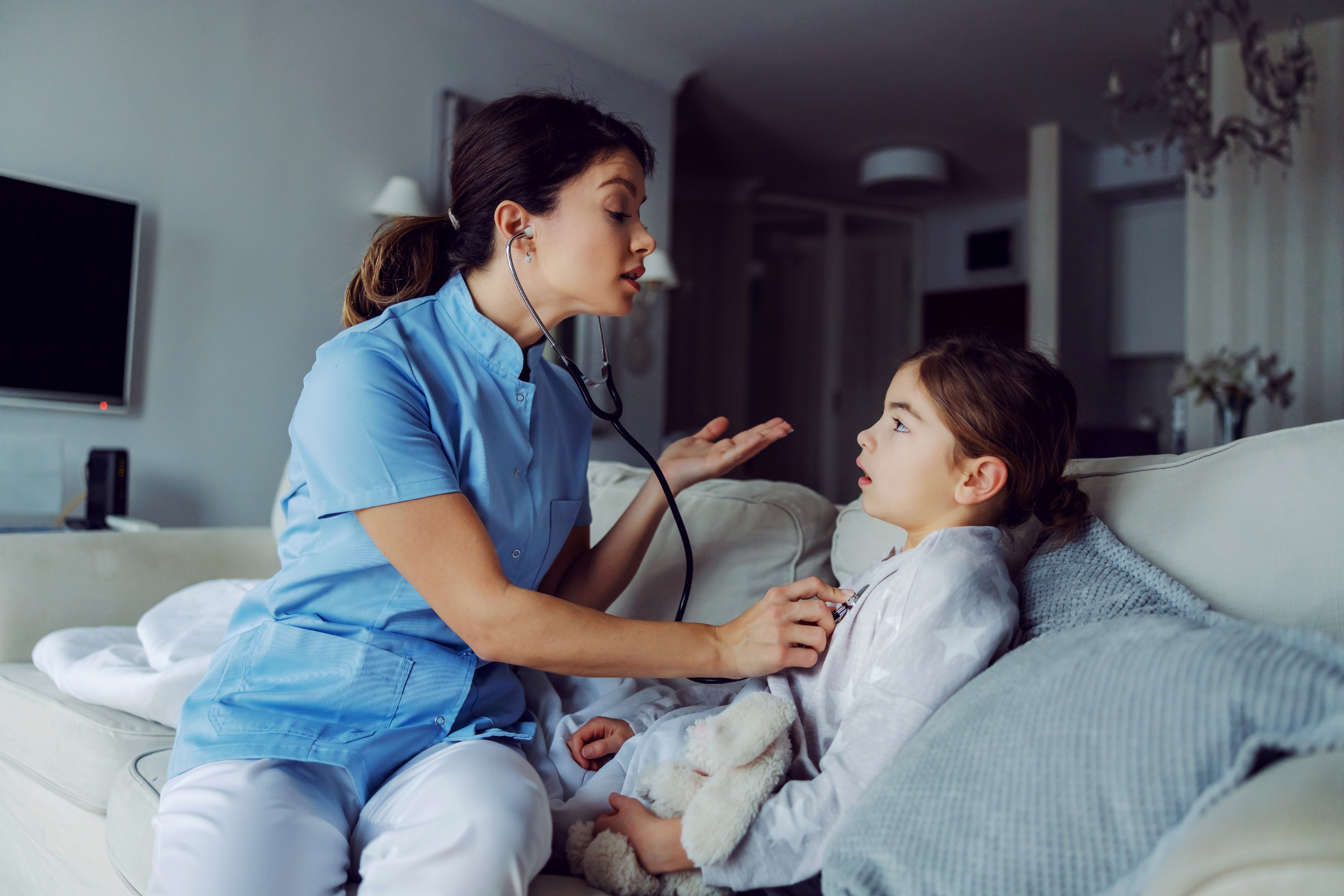 Médico sentado en un sofá junto a una niña y examinando sus pulmones con un estetoscopio | Fuente: Shutterstock.com