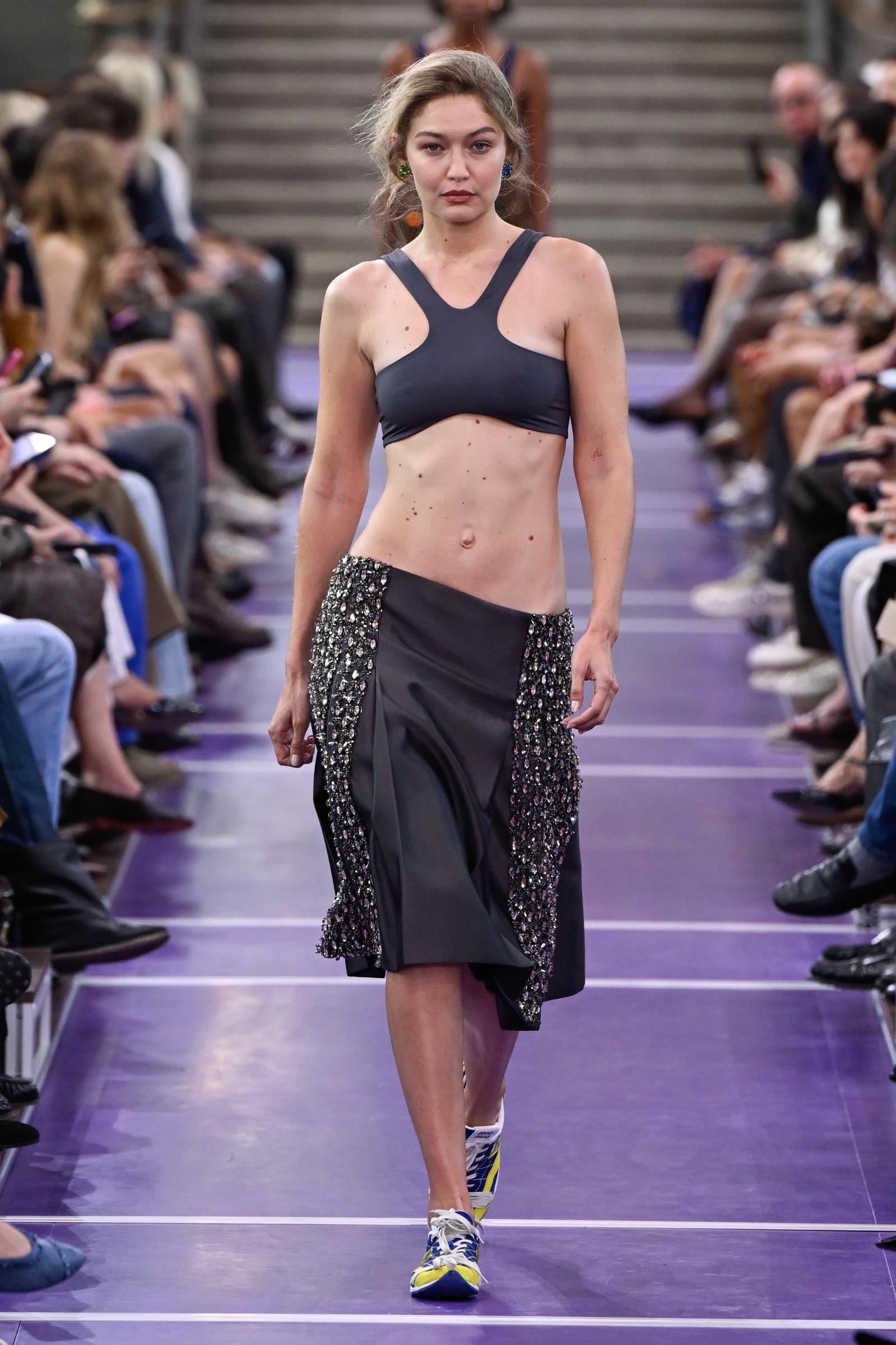Gigi Hadid arrasa en la pasarela de Miu Miu con un top corto en tono carbón y una falda midi drapeada a juego, y remata el look con unas zapatillas de colores y su melena rubia peinada hacia atrás de forma desenfadada. | Fuente: Getty Images