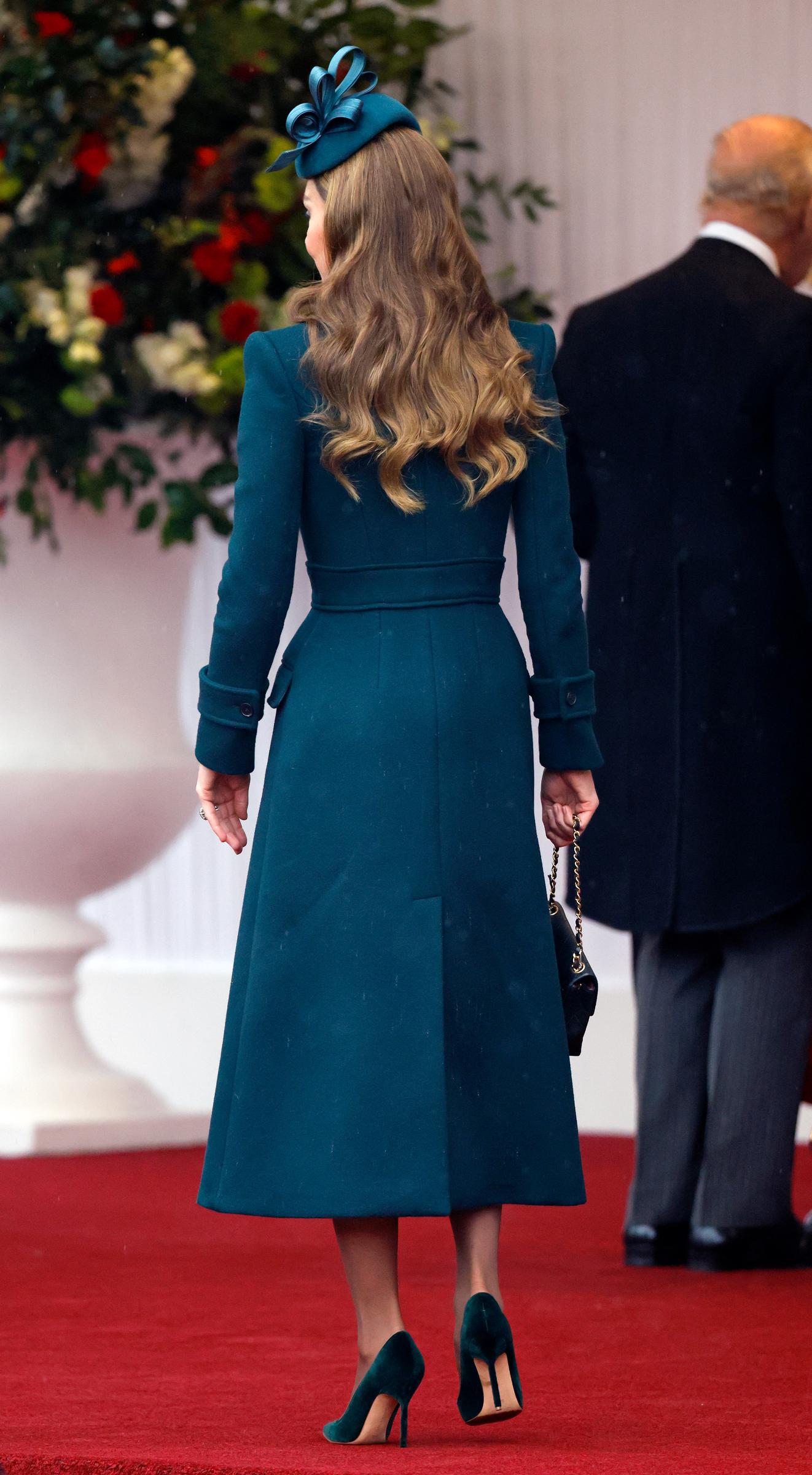 Una vista posterior de Catherine, princesa de Gales, resalta las líneas de su vestido-abrigo midi entallado mientras camina por la alfombra roja en Windsor el 20 de octubre de 2026. El conjunto verde oscuro luce cinturón ajustado y abertura central trasera. Su cabello castaño cae en suaves ondas bajo un tocado de Juliette Millinery. Completa el look con zapatos de tacón de aguja de terciopelo y un bolso Chanel acolchado que sostiene por su cadena. | Fuente: Getty Images