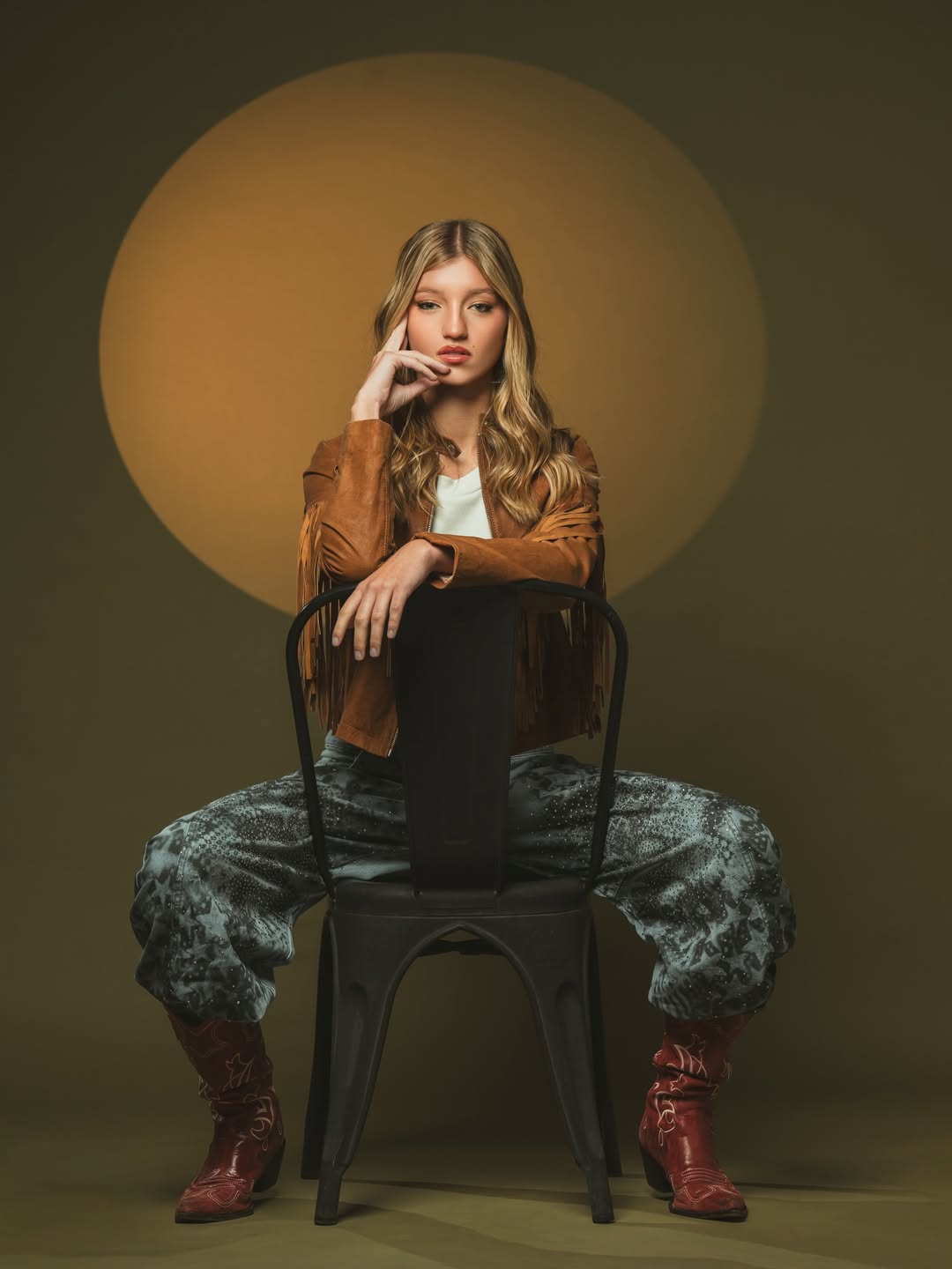Allegra Cubero posa sentada en una silla negra con una chaqueta marrón de flecos, pantalones estampados y botas de tono marrón rojizo | Foto: Lightpixel Producciones
