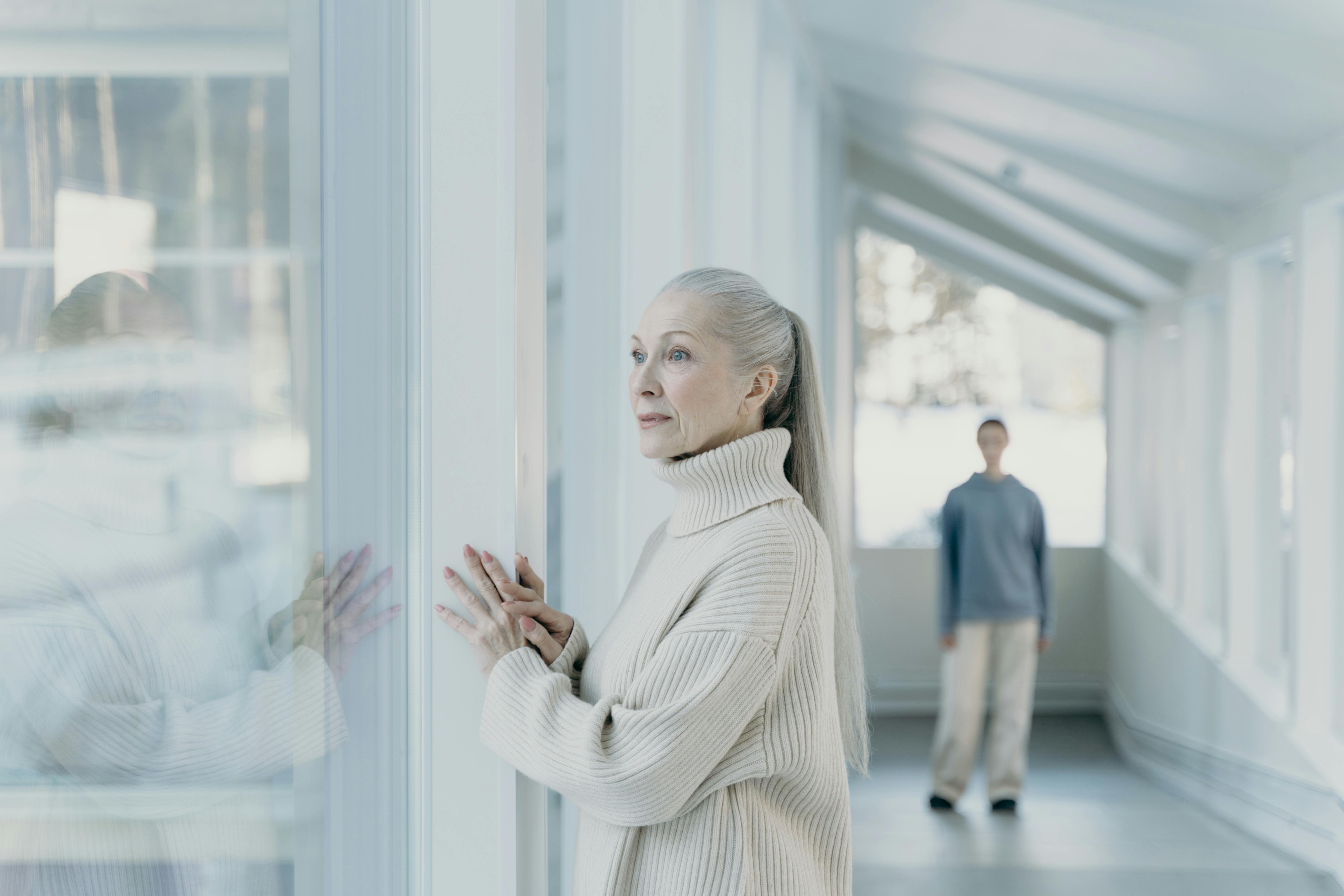 Una anciana mira a través del cristal de una ventana, con una figura al fondo | Fuente: Pexels