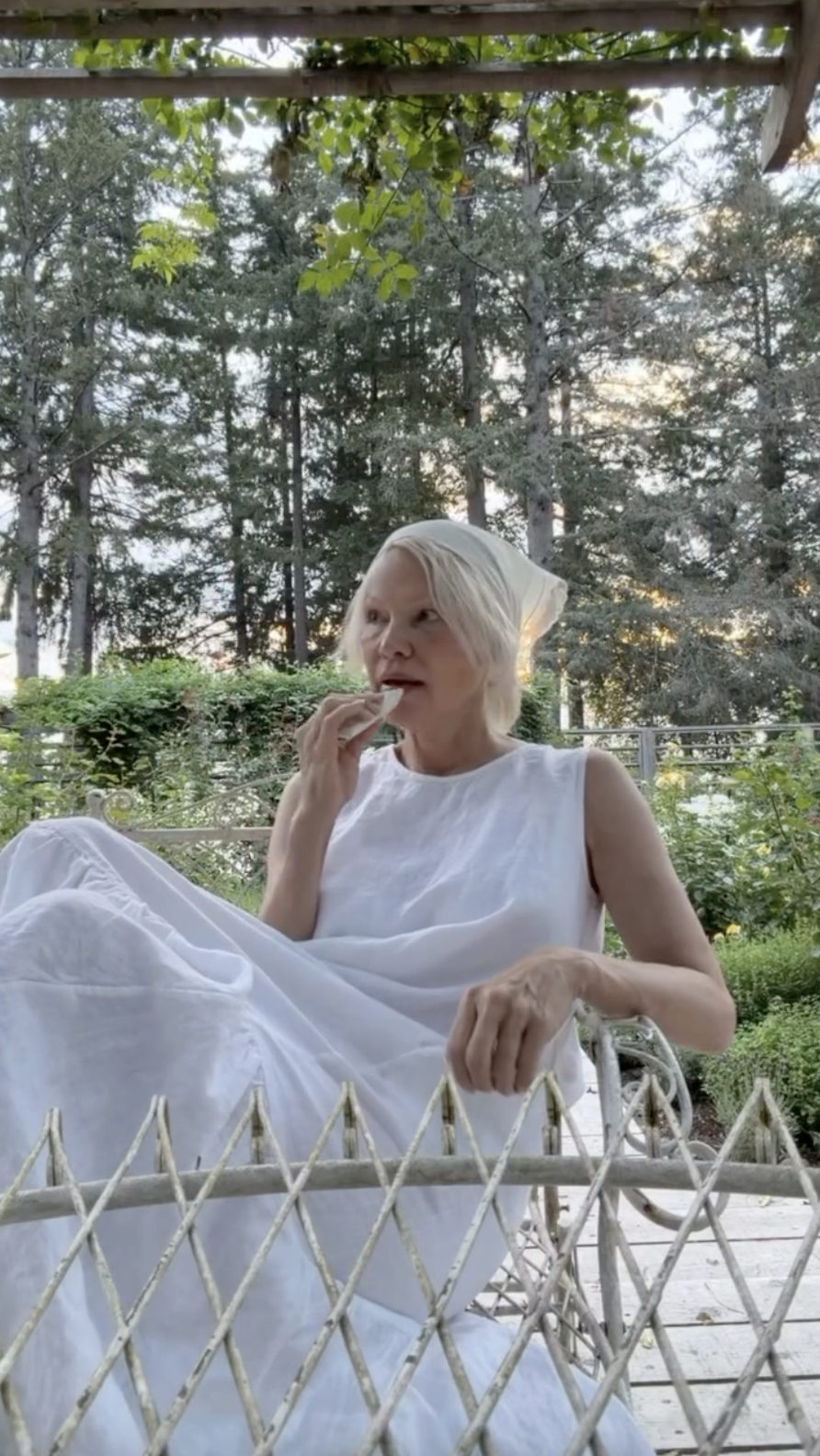 Pamela Anderson descansa tranquilamente en una silla de jardín vintage en Arcady, envuelta en un vestido blanco sin mangas con volantes y con un pañuelo color crema atado alrededor de su melena rubia. Con la mirada perdida en la lejanía mientras disfruta tranquilamente de algo para picar, encarna el ritmo tranquilo y relajado de la vida en la finca, escondida entre los altos pinos de Columbia Británica, y comparte el momento con el siguiente pie de foto: "Vive con romanticismo estés donde estés… 🤍🌷". | Fuente: Instagram/pamelaanderson