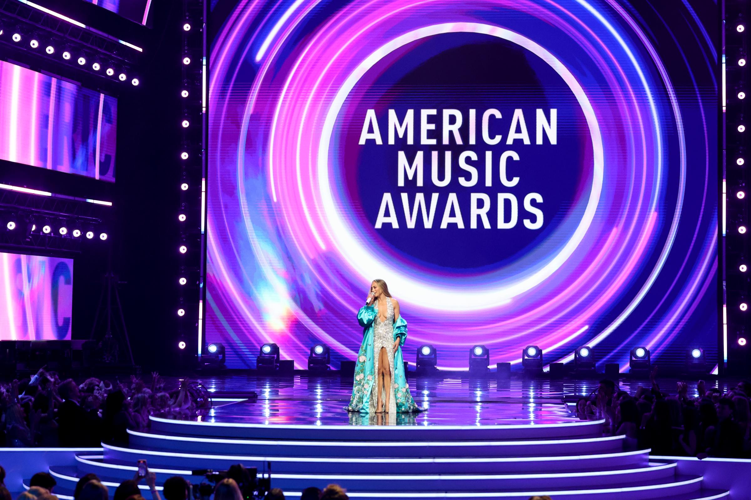 En una amplia toma del deslumbrante escenario, la cantante de 55 años se convierte en el centro de atención de los American Music Awards, enmarcada por un remolino de imágenes de neón ante la mirada del público.