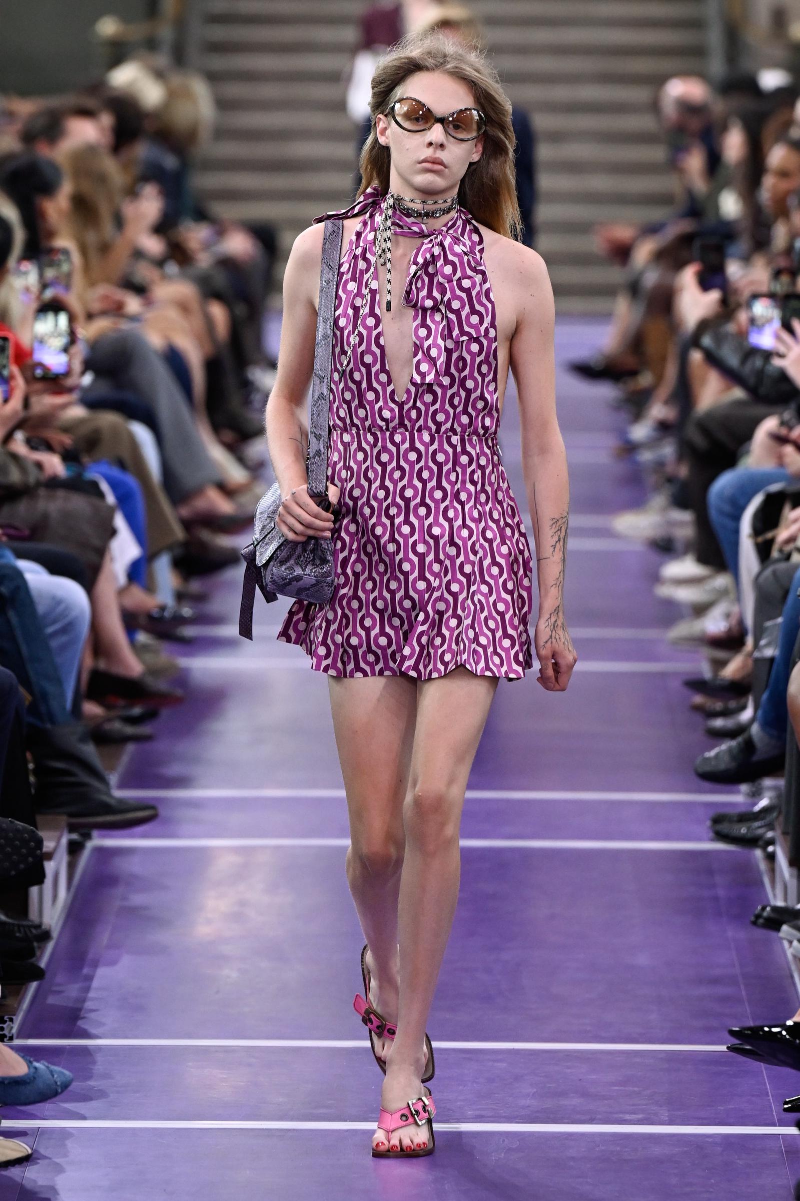 Una modelo cierra el desfile con un minivestido halter geométrico en tonos morados y blancos, gafas de sol extragrandes y gargantillas superpuestas, llevando un bolso de hombro metalizado y unos zapatos de tacón con tiras de color rosa. | Fuente: Getty Images