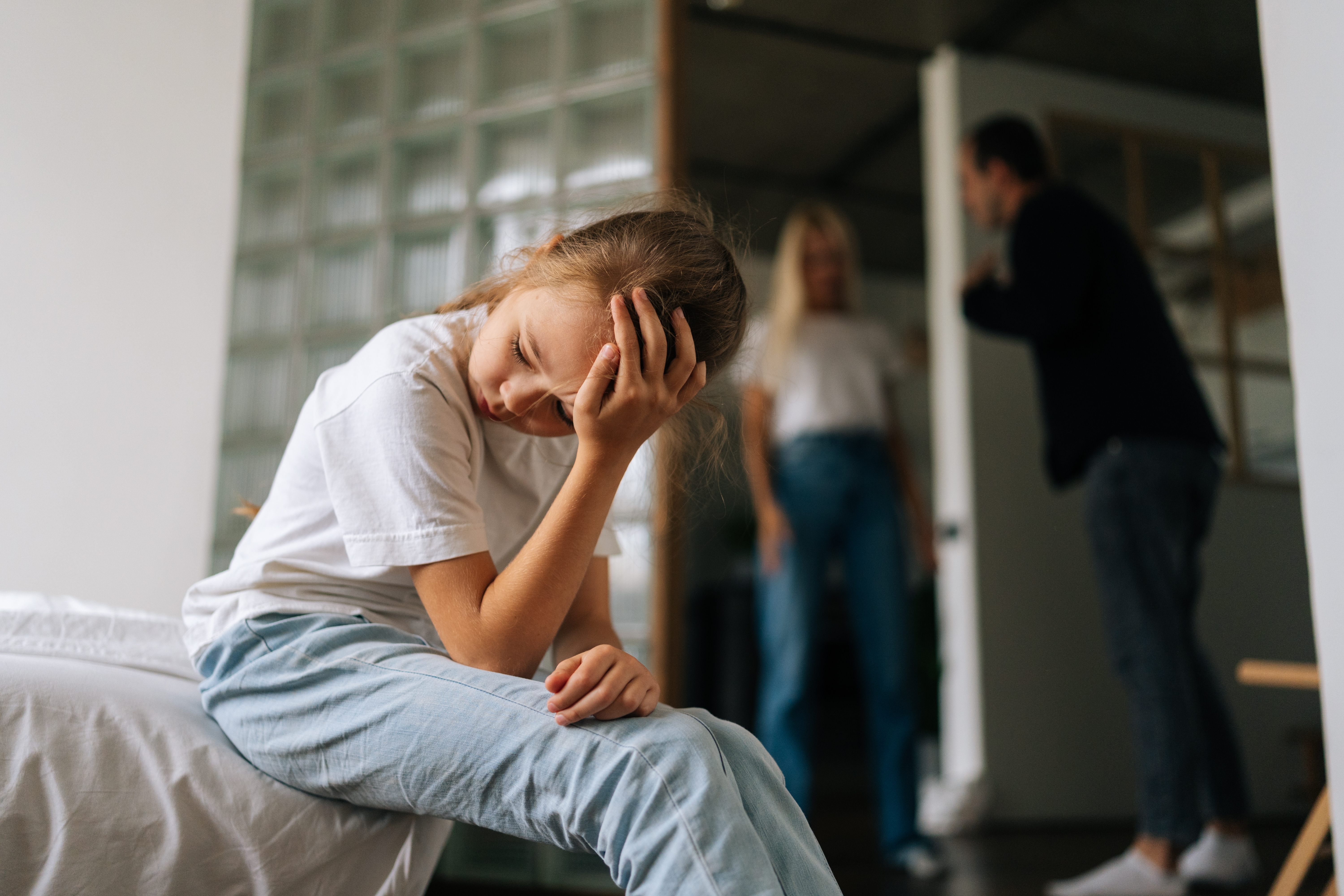 Una niña enfadada sentada en la esquina de su cama mientras sus padres discuten al fondo | Foto: Shutterstock