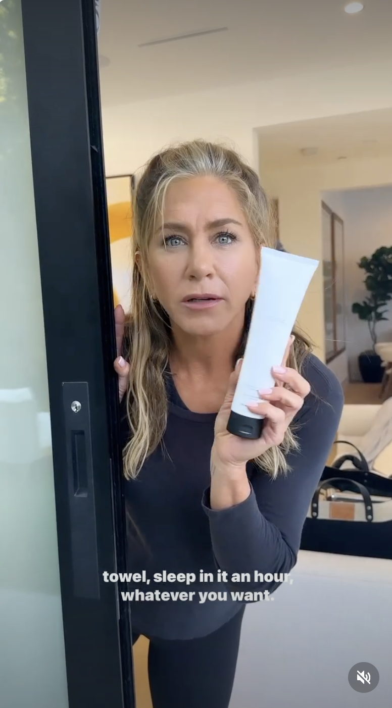Jennifer Aniston luce unas canas en la línea del pelo mientras sostiene un frasco del tratamiento reparador intensivo de LolaVie en un video de Instagram de junio de 2023. La actriz lleva una camiseta oscura de manga larga y el pelo rubio recogido a medias. | Fuente: Instagram/jenniferaniston