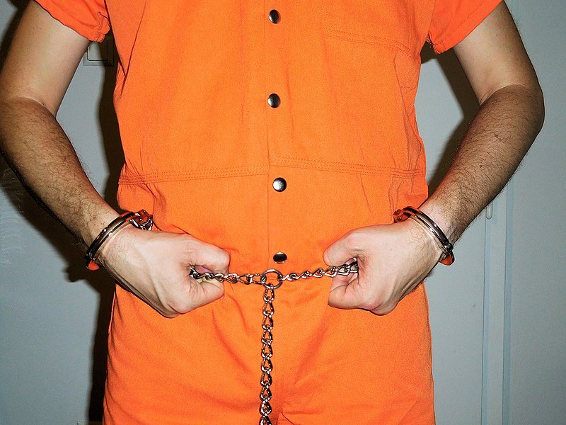 Primer plano de un recluso en bata institucional naranja y restricciones de transporte de "arnés completo". | Imagen: Wikimedia Commons