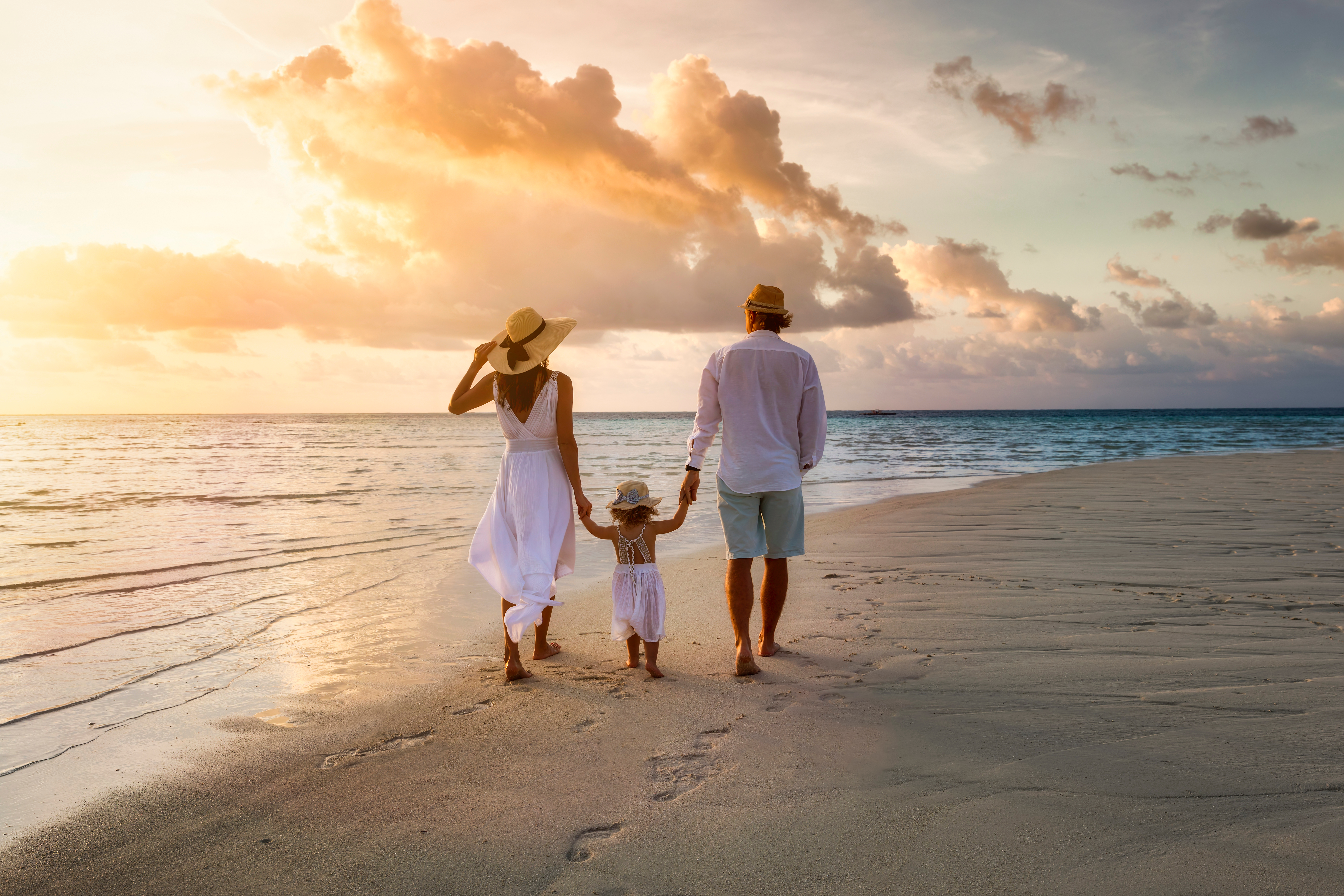 Una elegante familia con ropa blanca de verano paseando de la mano por una playa paradisíaca tropical durante la puesta de sol | Foto: Shutterstock