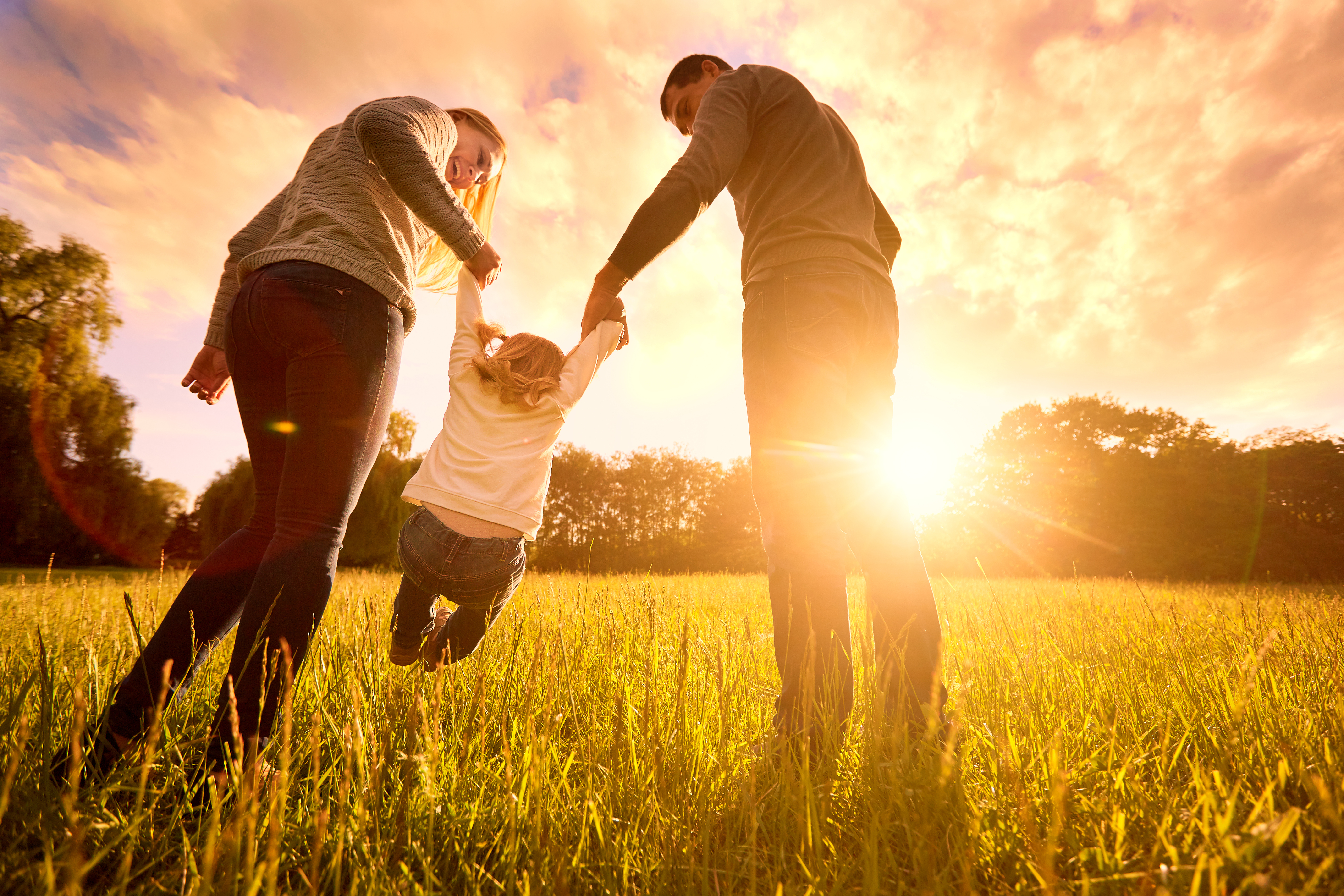 Un hombre y una mujer levantan a un niño en el aire sobre un campo de hierba con la puesta de sol como telón de fondo | Fuente: Shutterstock