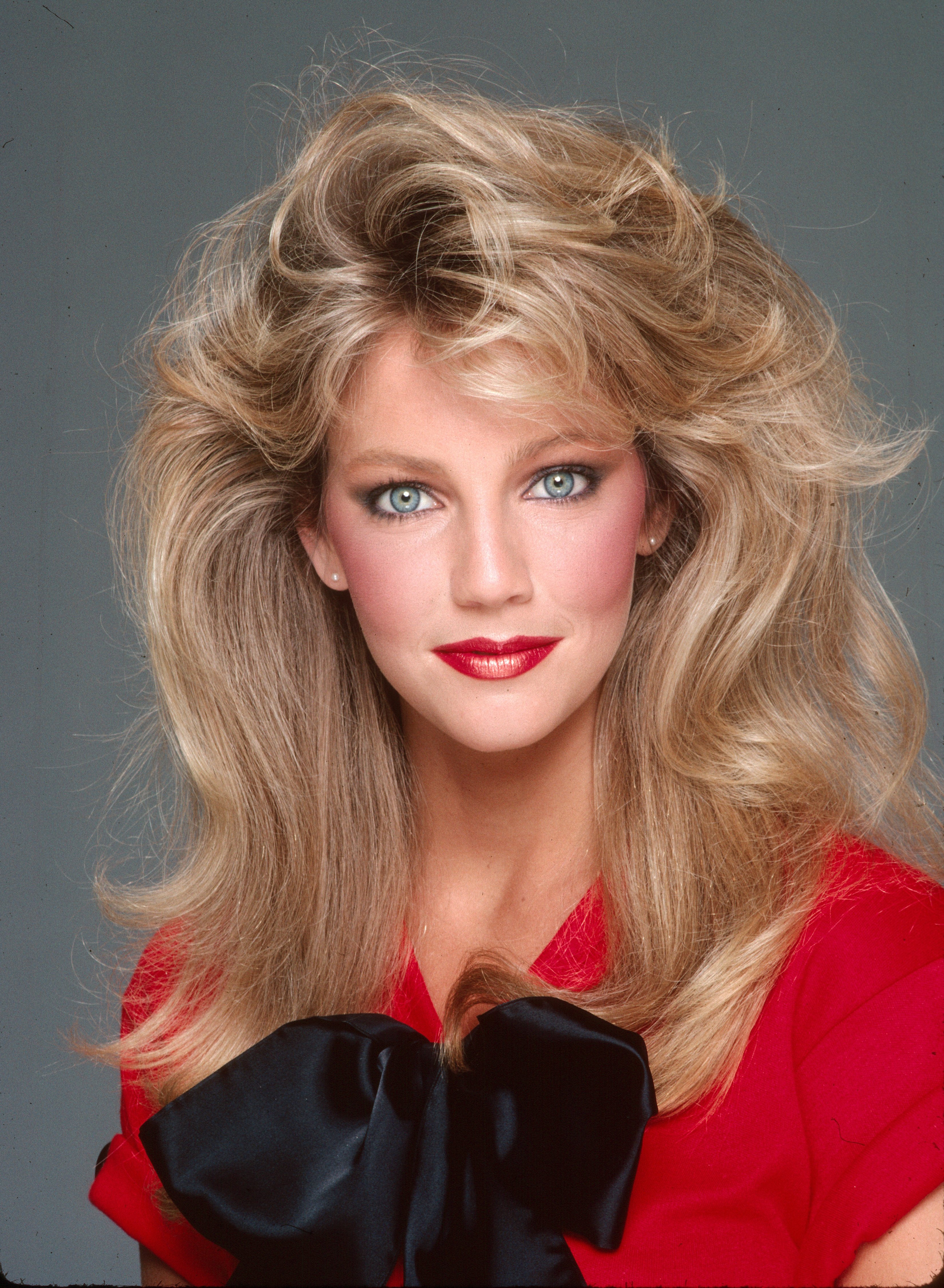 Heather Locklear es el paradigma del glamour de los años 80 en una sesión de fotos de estudio del 2 de febrero de 1981 en Los Ángeles, California. Con unas voluminosas ondas rubias en capas que le enmarcan el rostro, mira directamente a la cámara luciendo un rubor rosado, un labial rojo intenso y unos aretes de perla, mientras lleva un top rojo vivo adornado con un lazo extragrande de satén negro. | Fuente: Getty Images