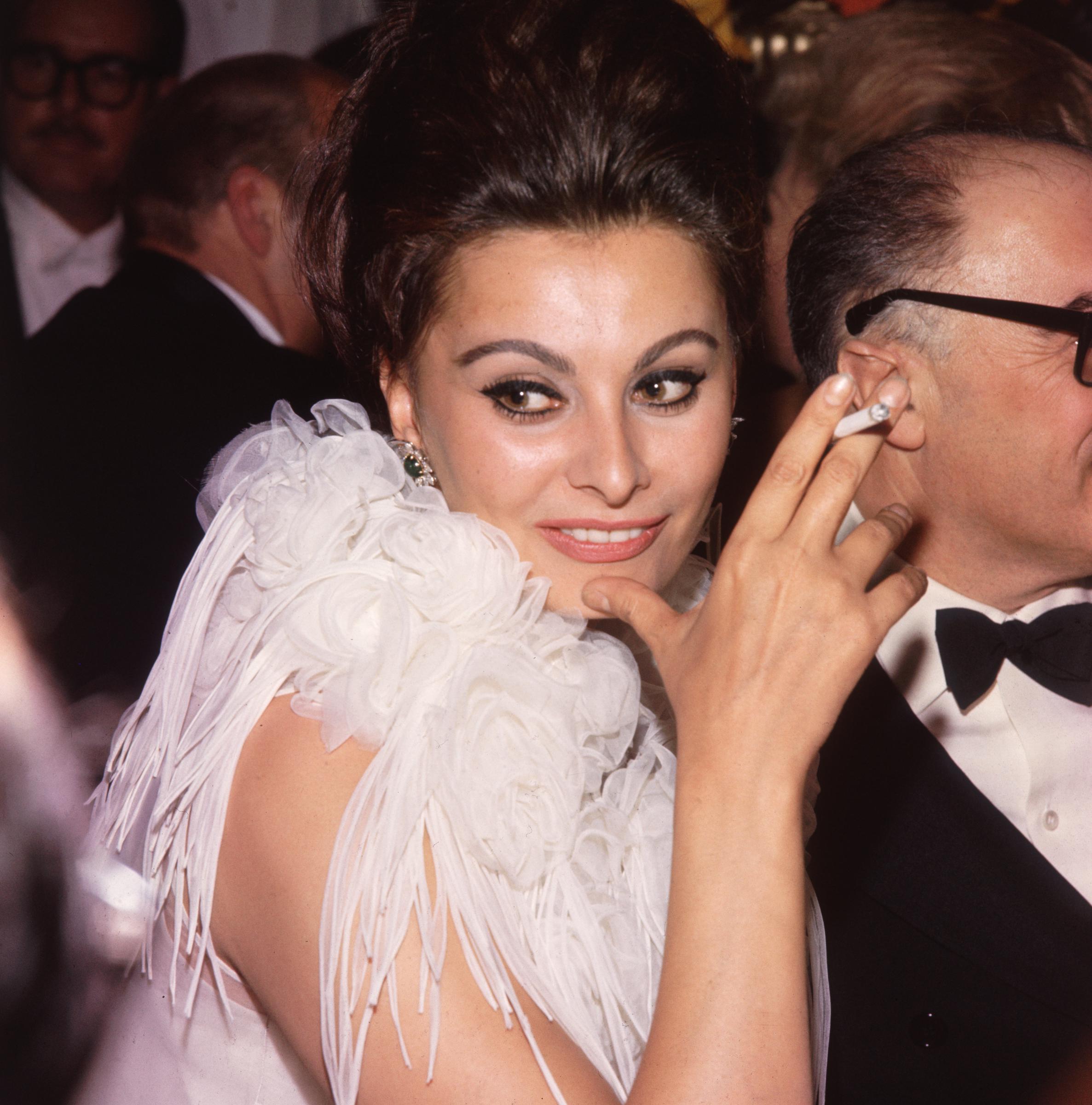 Foto de la actriz italiana Sophia Loren, vestida con un traje con cuello de roseta de gasa blanca, fumando un cigarrillo en los Premios de la Academia, Los Ángeles, California. | Fuente: Getty Images.