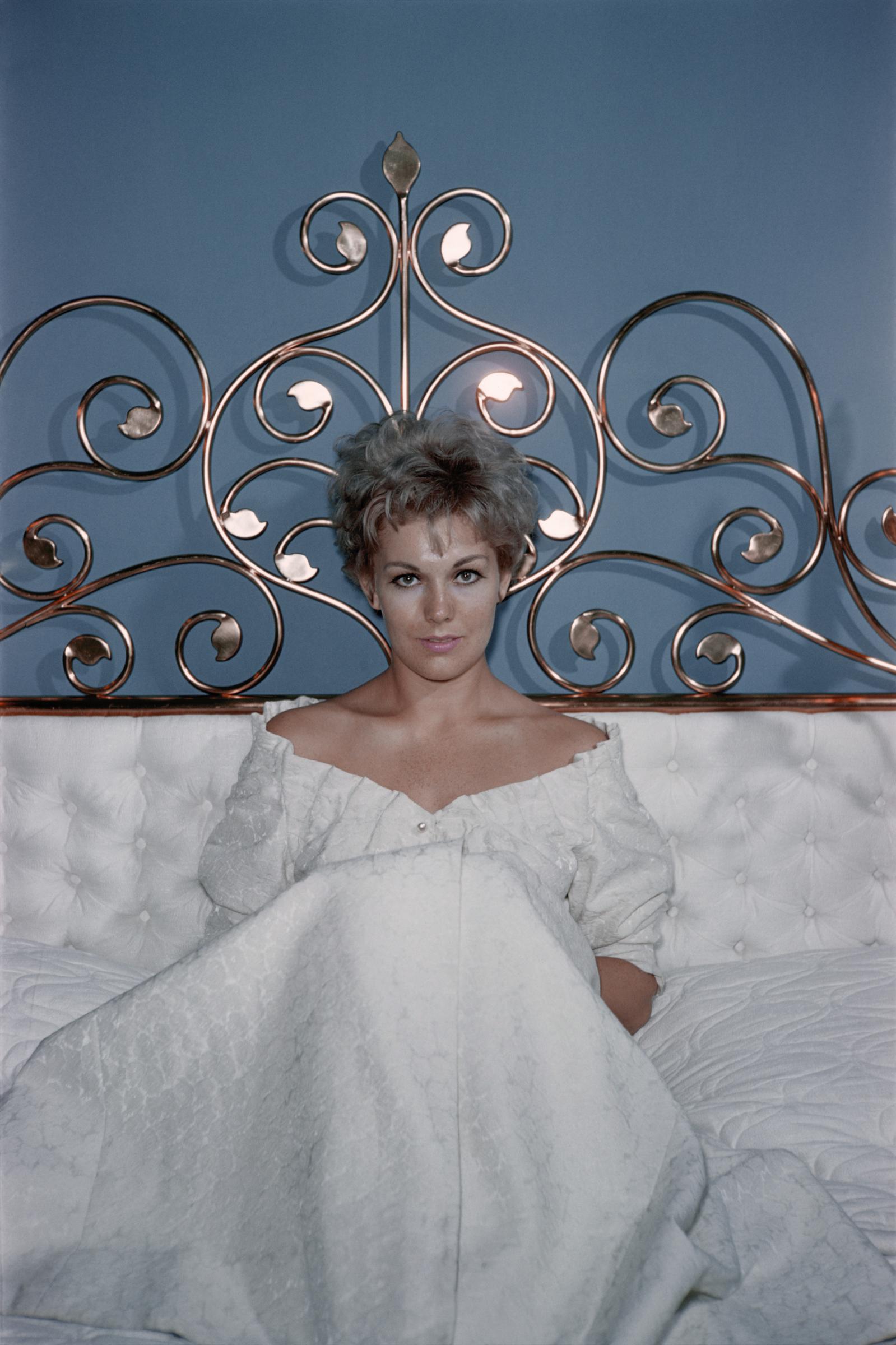Kim Novak está sentada en una cama con un vestido blanco con los hombros al aire, posando frente a un cabecero metálico muy decorado. | Fuente: Getty Images