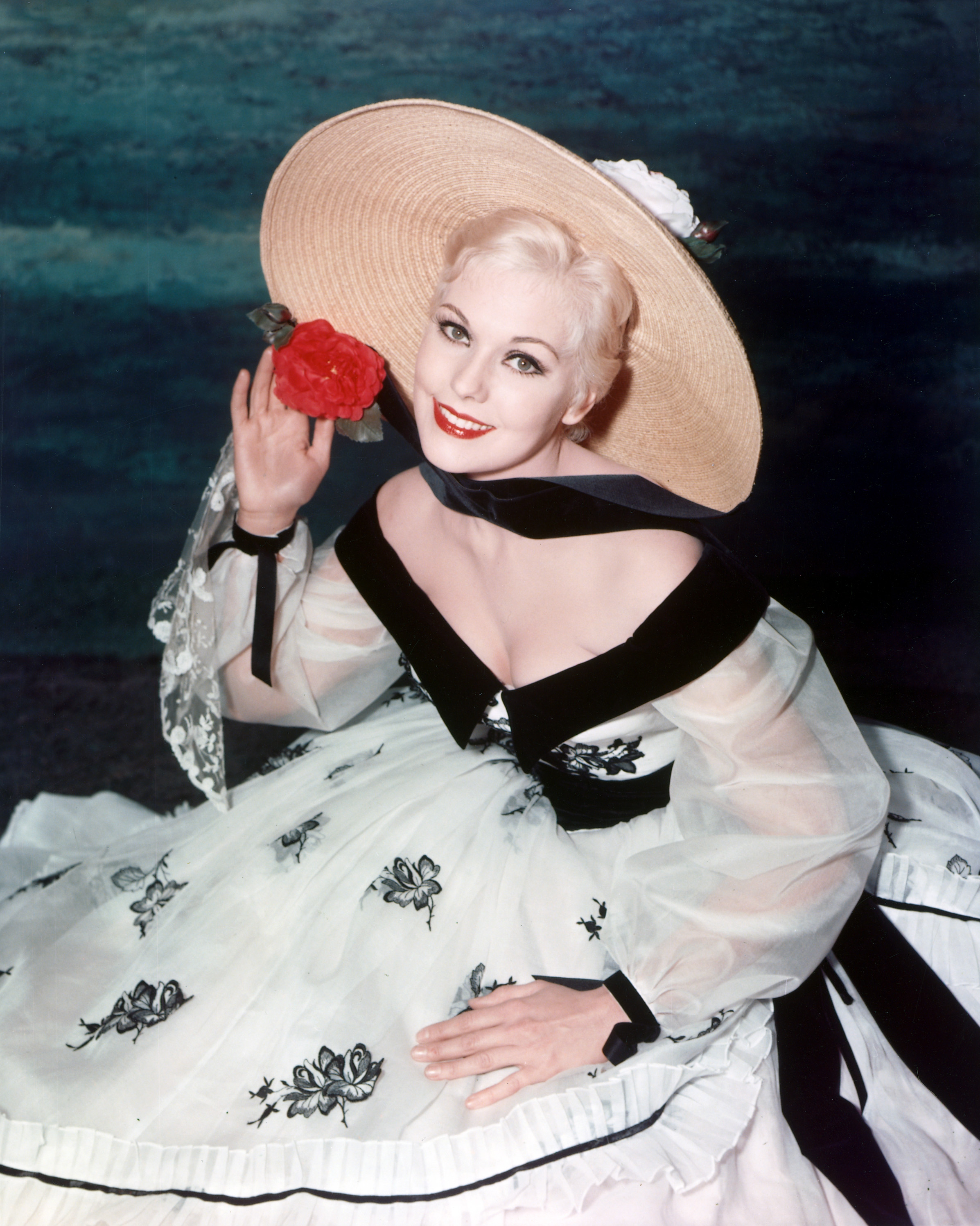 Kim Novak sonríe en un retrato de estudio con un vestido blanco con detalles florales negros y un sombrero de paja de ala ancha adornado con una flor roja. | Fuente: Getty Images