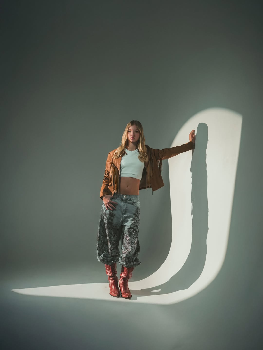 Allegra Cubero posa de pie apoyando una mano en una pared, vistiendo una chaqueta marrón de flecos, un top corto blanco, pantalones estampados y botas de tono marrón rojizo | Foto: Lightpixel Producciones