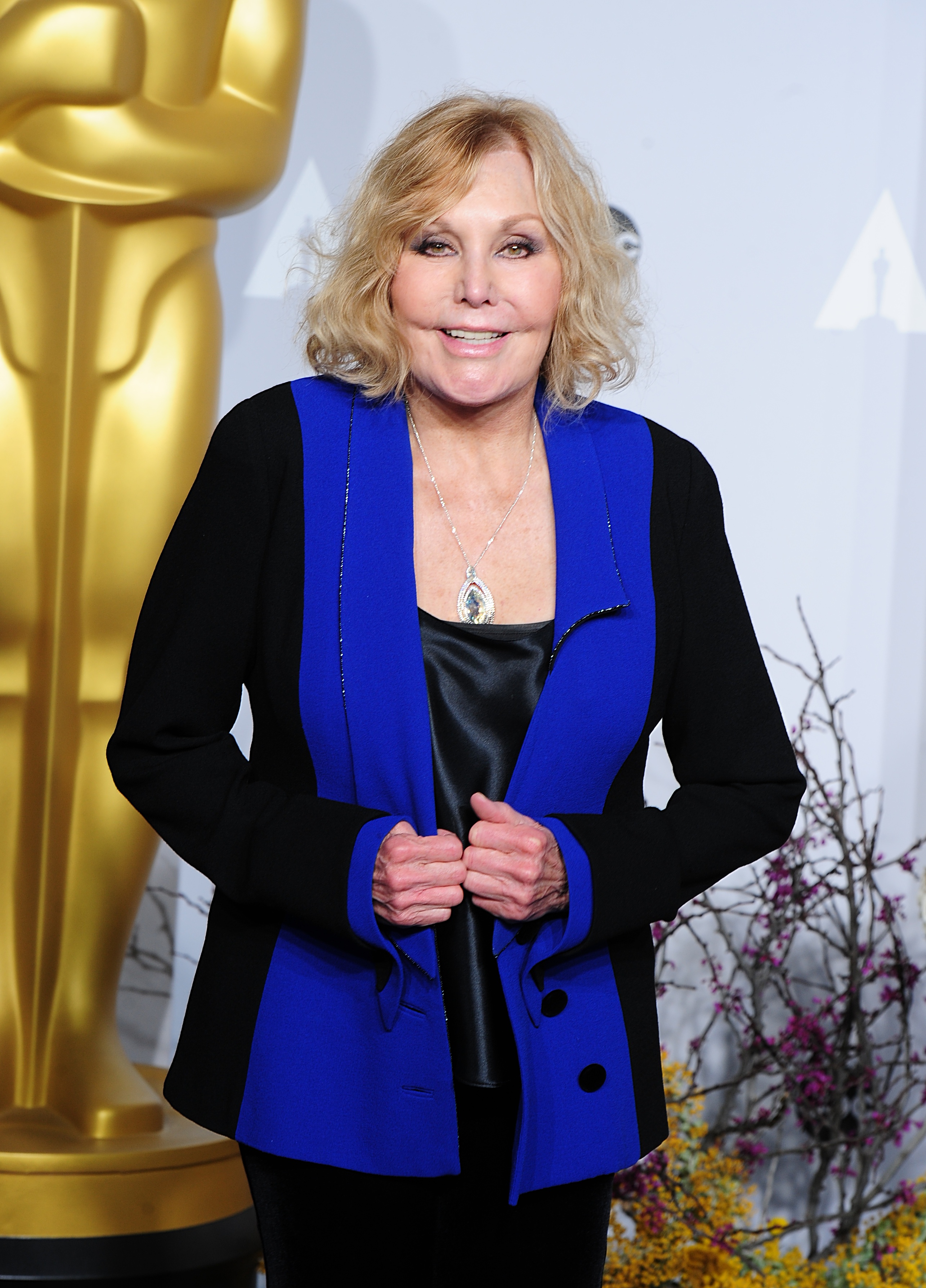 Kim Novak sonríe a los fotógrafos con un conjunto negro con solapas de un azul intenso en la 86.ª edición de los Premios de la Academia, celebrada en Hollywood el 2 de marzo de 2014. | Fuente: Getty Images