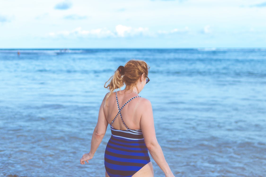 Mujer utilizando un traje de baño entero de rayas azules y negras con tirantes mientras camina en la playa. | Foto: Pexels