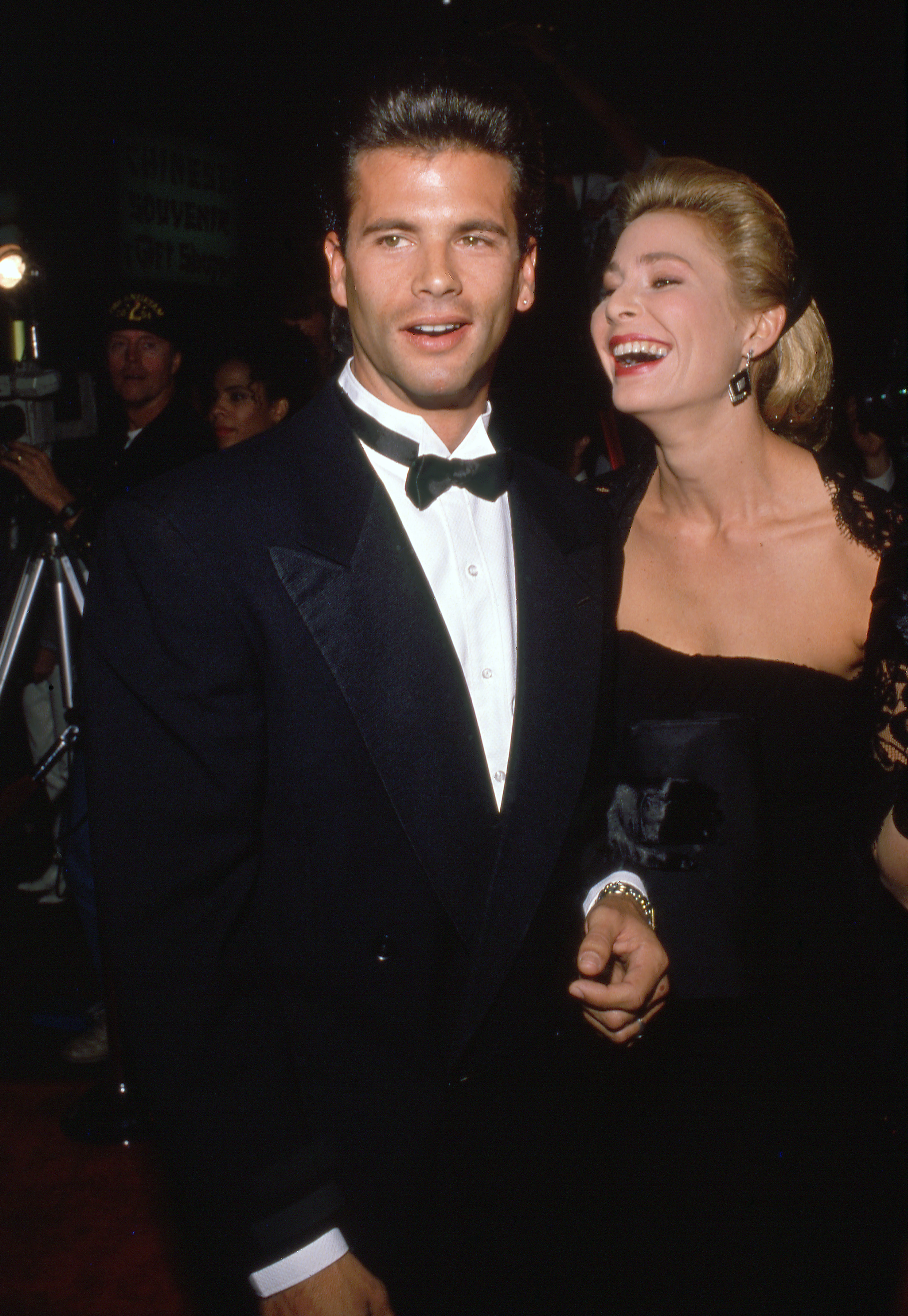 Lorenzo Lamas y su entonces esposa, la actriz Kathleen Kinmont, disfrutan de un momento alegre en la alfombra roja durante un evento nocturno de los años 90. Lamas luce impecable con un esmoquin negro clásico, una camisa blanca plisada y un corbatín negro, mientras que Kinmont se ríe a su lado con un elegante vestido negro, el pelo rubio peinado hacia atrás y unos aretes llamativos. | Fuente: Getty Images