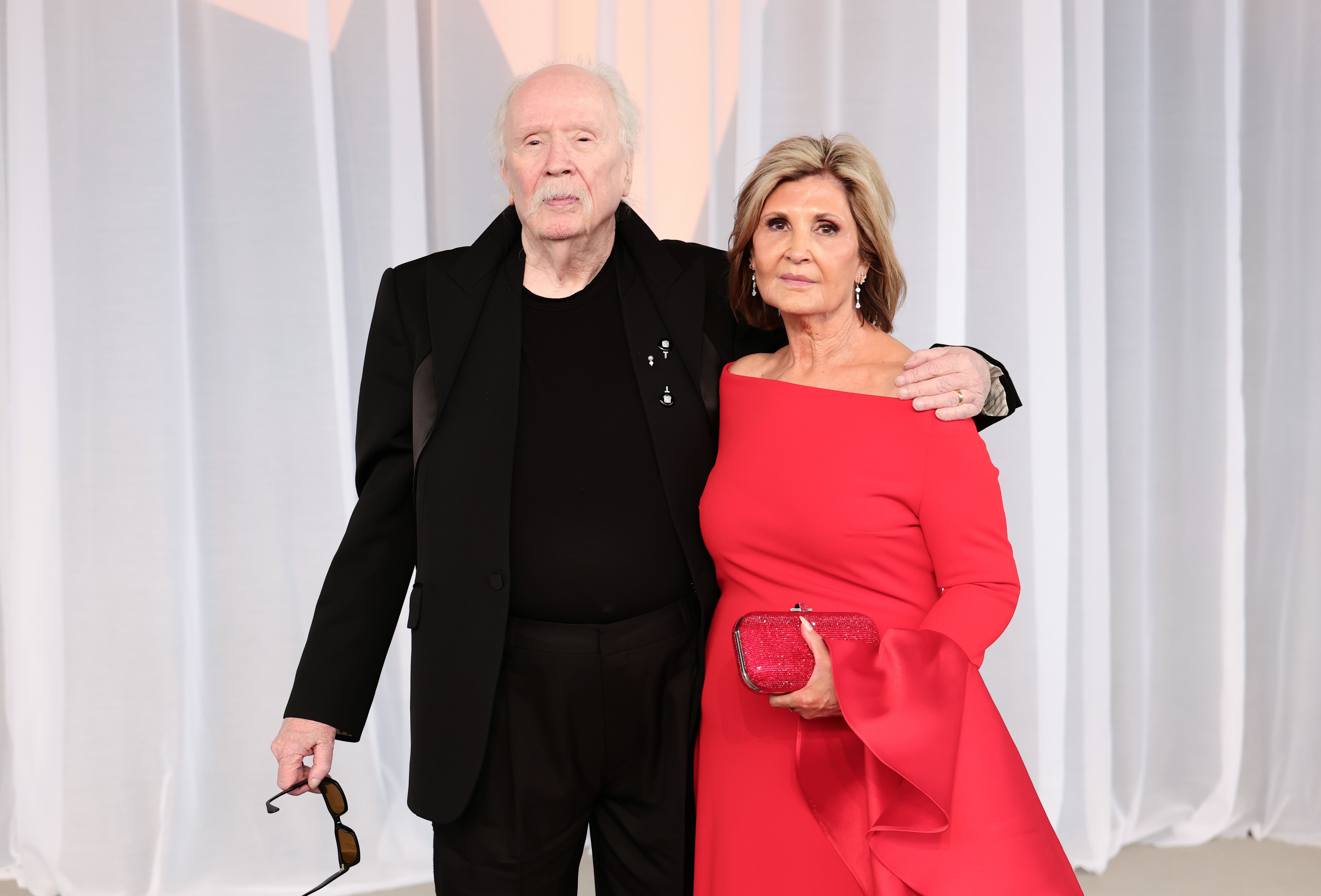 John Carpenter y su esposa, la productora Sandy King, posan juntos con un look a juego. King destaca con un vestido rojo cereza de hombros al aire con unas espectaculares mangas acampanadas, que combina con un brillante bolso de mano color rubí, mientras que Carpenter opta por un traje a medida totalmente negro con solapas marcadas y un suéter de cuello redondo oscuro a juego, sosteniendo sus gafas de sol a un lado mientras celebra haber recibido el Premio Luminary.