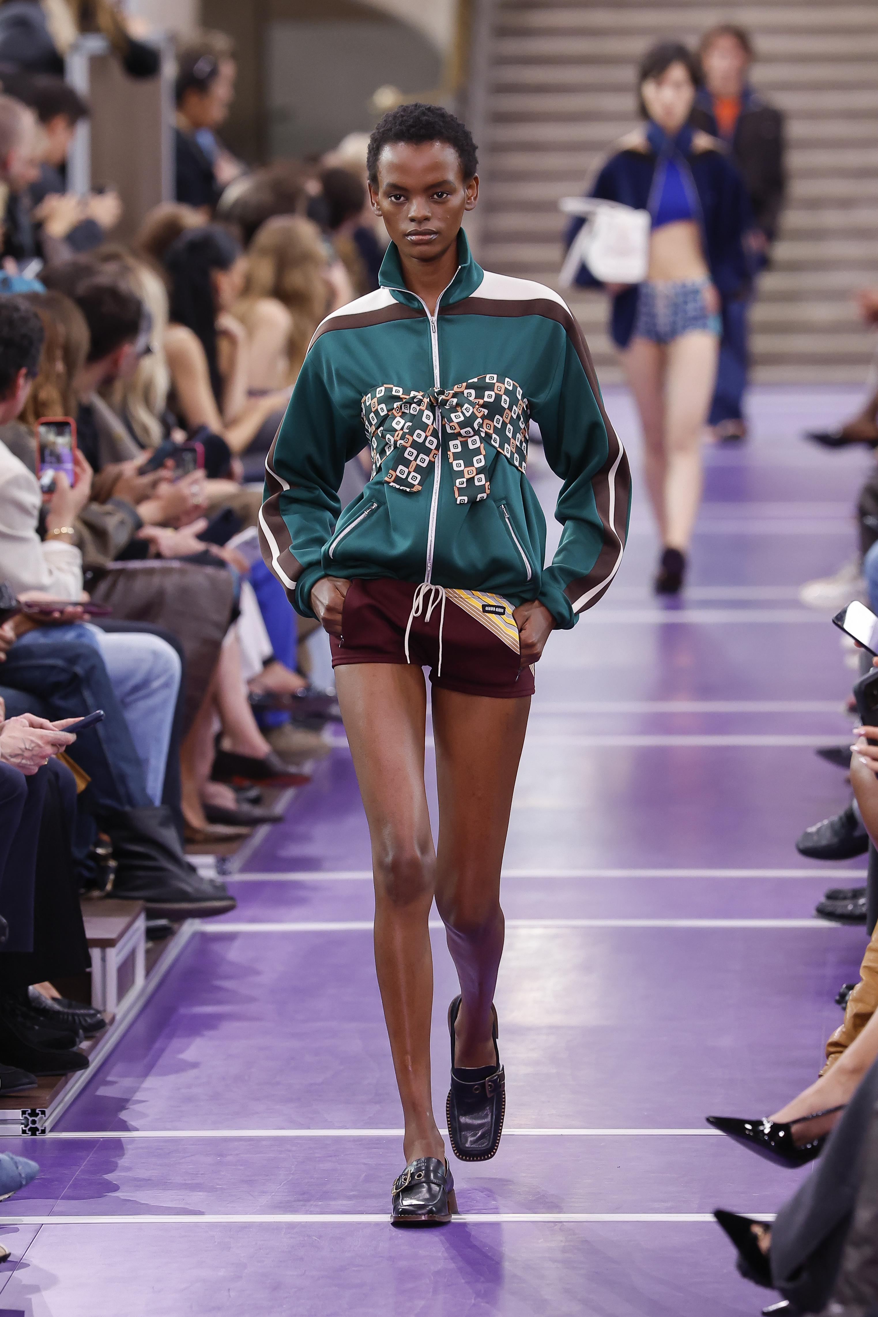 Una modelo desfila por la pasarela de Miu Miu con una chaqueta deportiva de color verde azulado con rayas inspiradas en el mundo del automovilismo y un lazo estampado, combinada con unos pantalones cortos burdeos y unos mocasines negros. | Fuente: Getty Images
