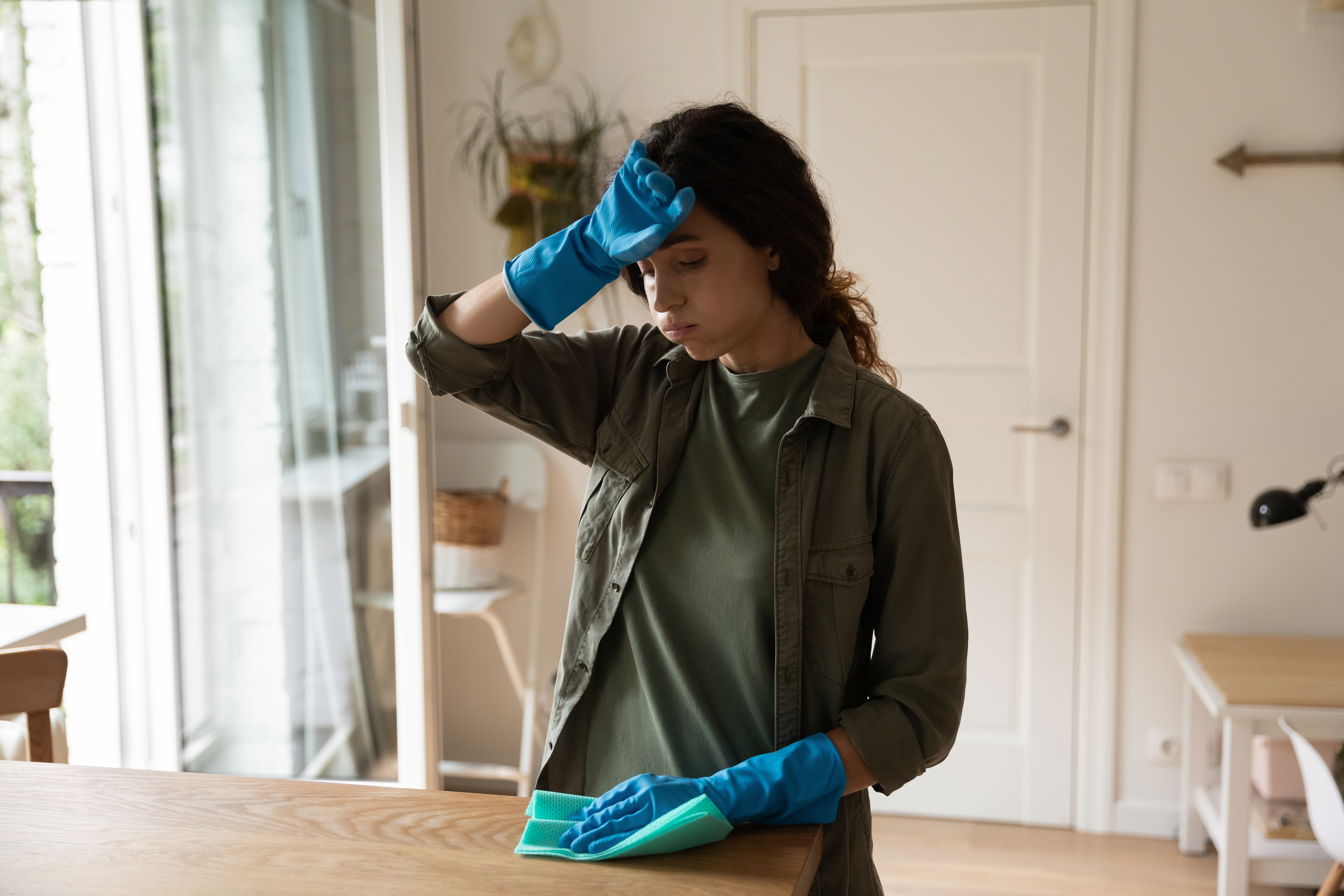 Exhausta ama de casa milenaria con guantes de látex azules se limpia el sudor de la frente. | Fuente: Shutterstock