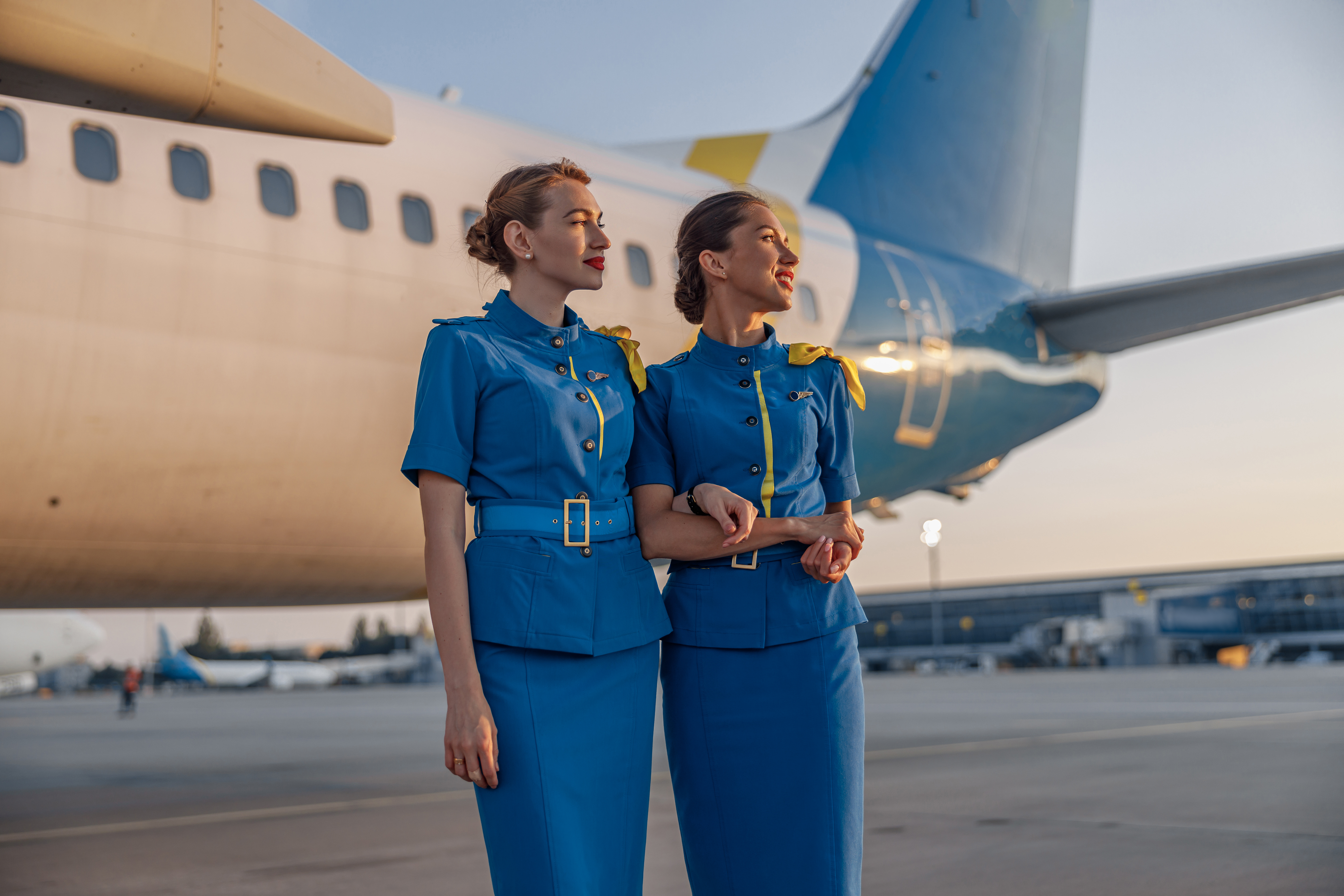 Dos hermosas azafatas con uniforme azul sonriendo, de pie delante de un gran avión de pasajeros en un aeropuerto al atardecer. | Fuente: Shutterstock