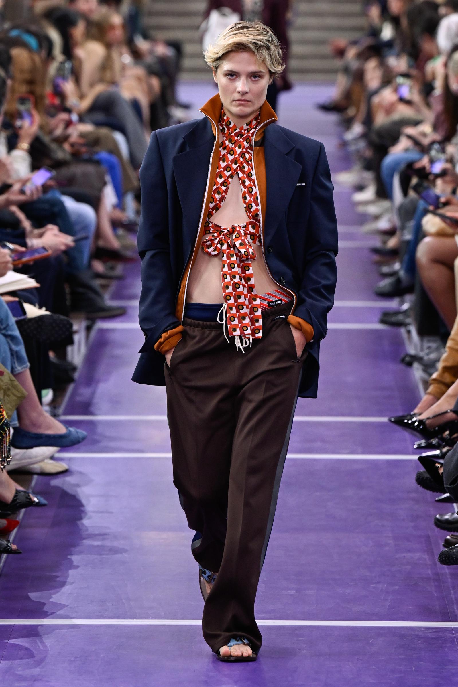 Louisa Jacobson desfila por la pasarela de Miu Miu con una chaqueta azul marino entallada con detalles naranjas a contraste sobre un top halter de estampado llamativo, combinado con unos pantalones marrones holgados y unas sandalias planas con la punta abierta. | Fuente: Getty Images