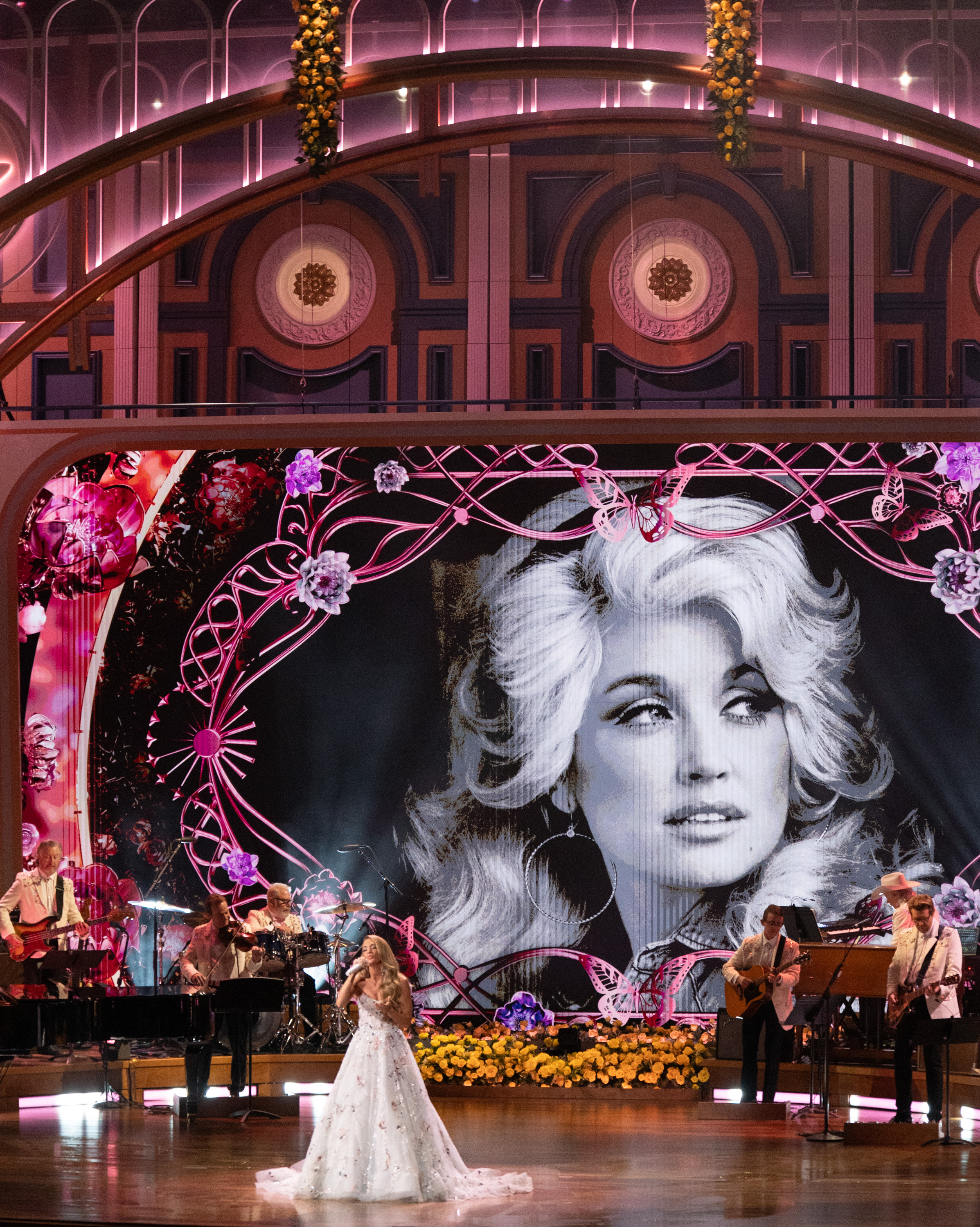 Carrie Underwood actúa bajo un retrato gigante en blanco y negro de Dolly Parton, rodeada de mariposas rosas y adornos florales durante el homenaje celebrado en Nashville. | Fuente: Getty Images.