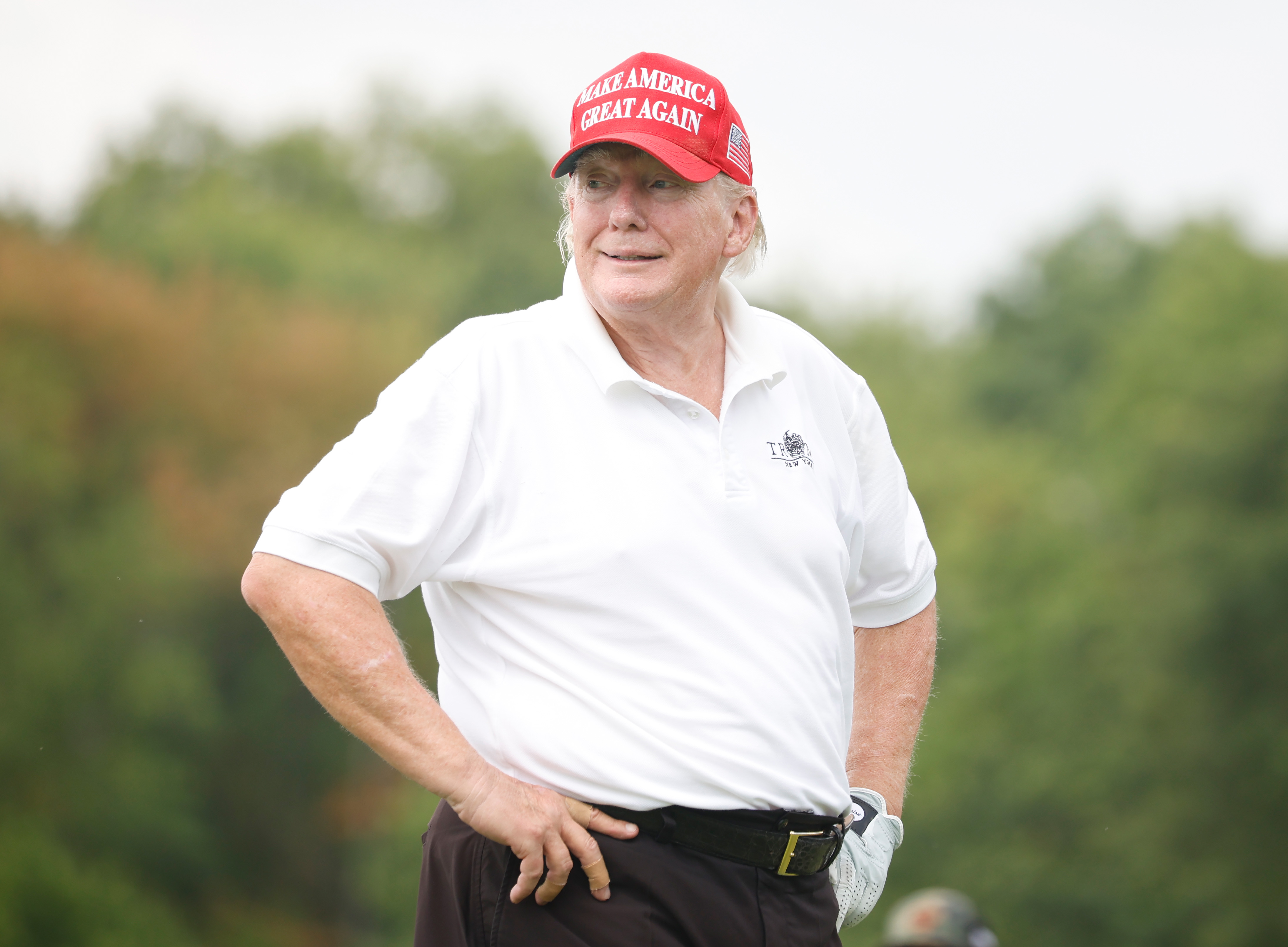 Con su característica gorra roja y un polo blanco, Donald Trump está de pie en el campo de golf con las manos en la cadera. Sus mejillas y su mandíbula aparecen notablemente enrojecidas, en contraste con la piel pálida de su cuello.
