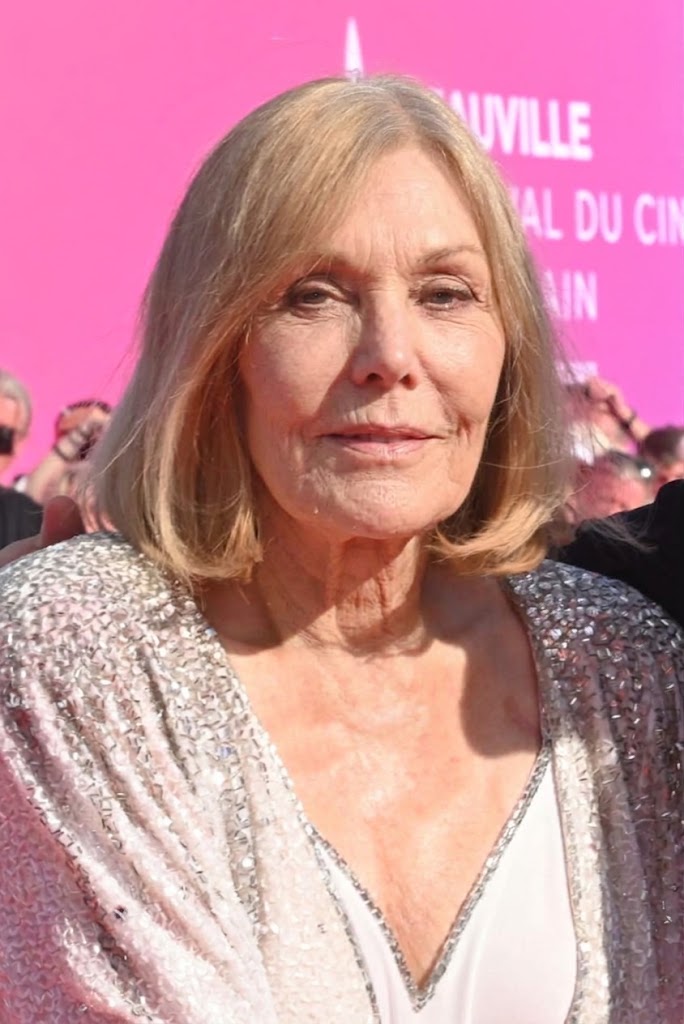 Kim Novak posa con ambas manos apoyadas cerca del pecho, con un vestido blanco de cuello en V y una chaqueta plateada brillante, mientras un telón de fondo rosa enmarca el retrato. | Fuente: Gemini