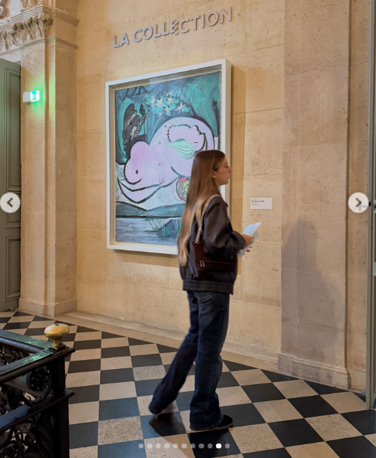 Antonia Macri recorre una galería de arte con paso tranquilo sobre un piso ajedrezado, luciendo jeans acampanados, campera oscura y una cartera al hombro con cadena mientras contempla una pintura en exhibición. | Fuente: Instagram/juliana.awada