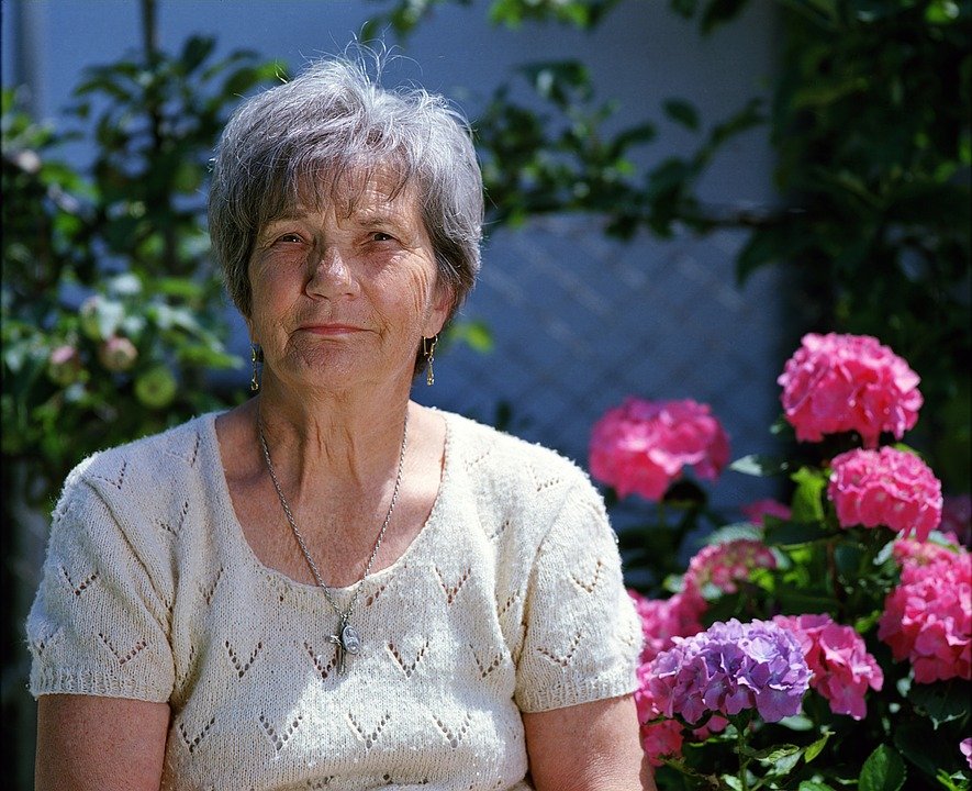 Mujer mayor sentada frente a un jardín de flores. │Foto: Pixabay
