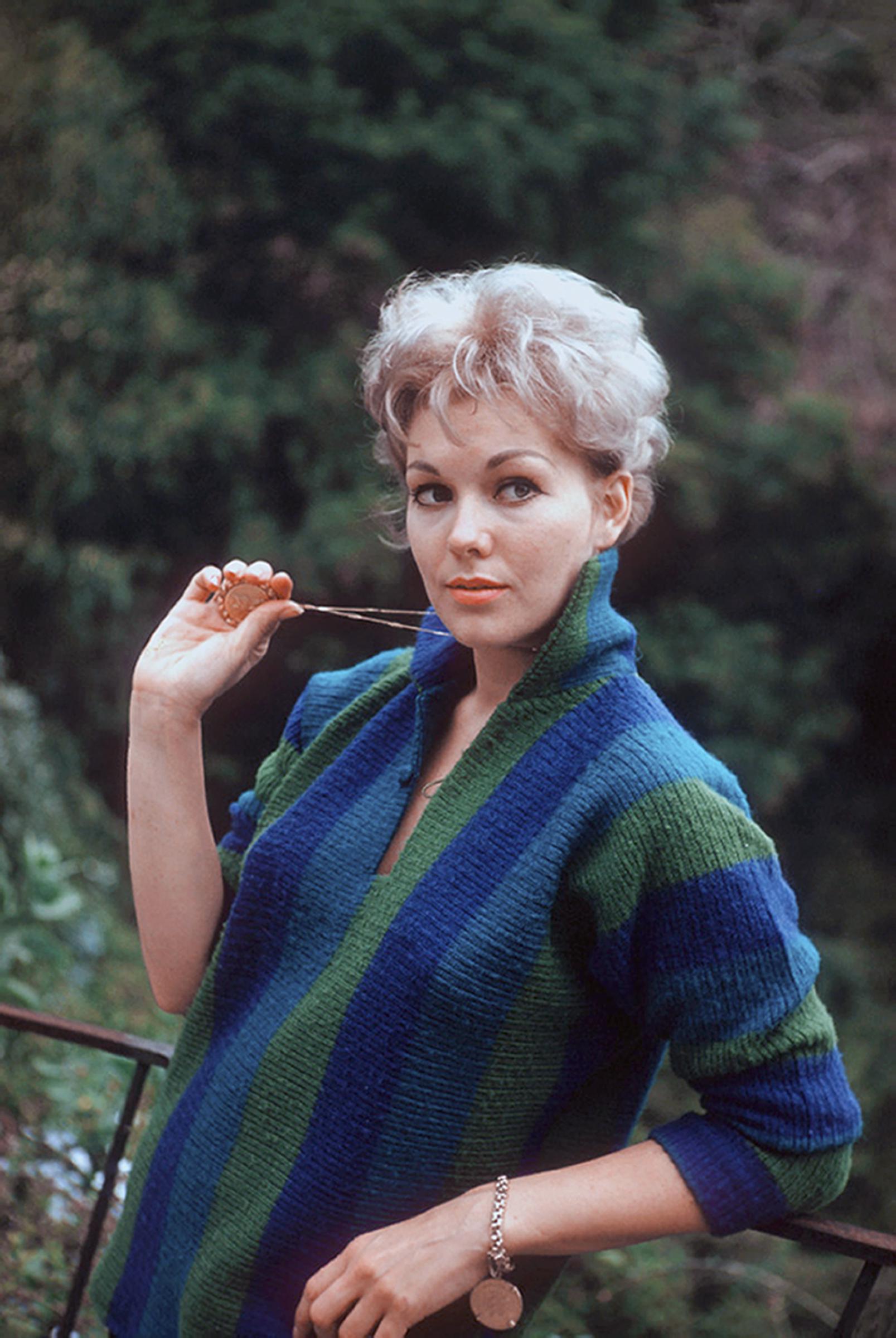 Kim Novak posa en su casa con un colorido conjunto de punto azul y verde, sosteniendo un colgante cerca de la cara. | Fuente: Getty Images