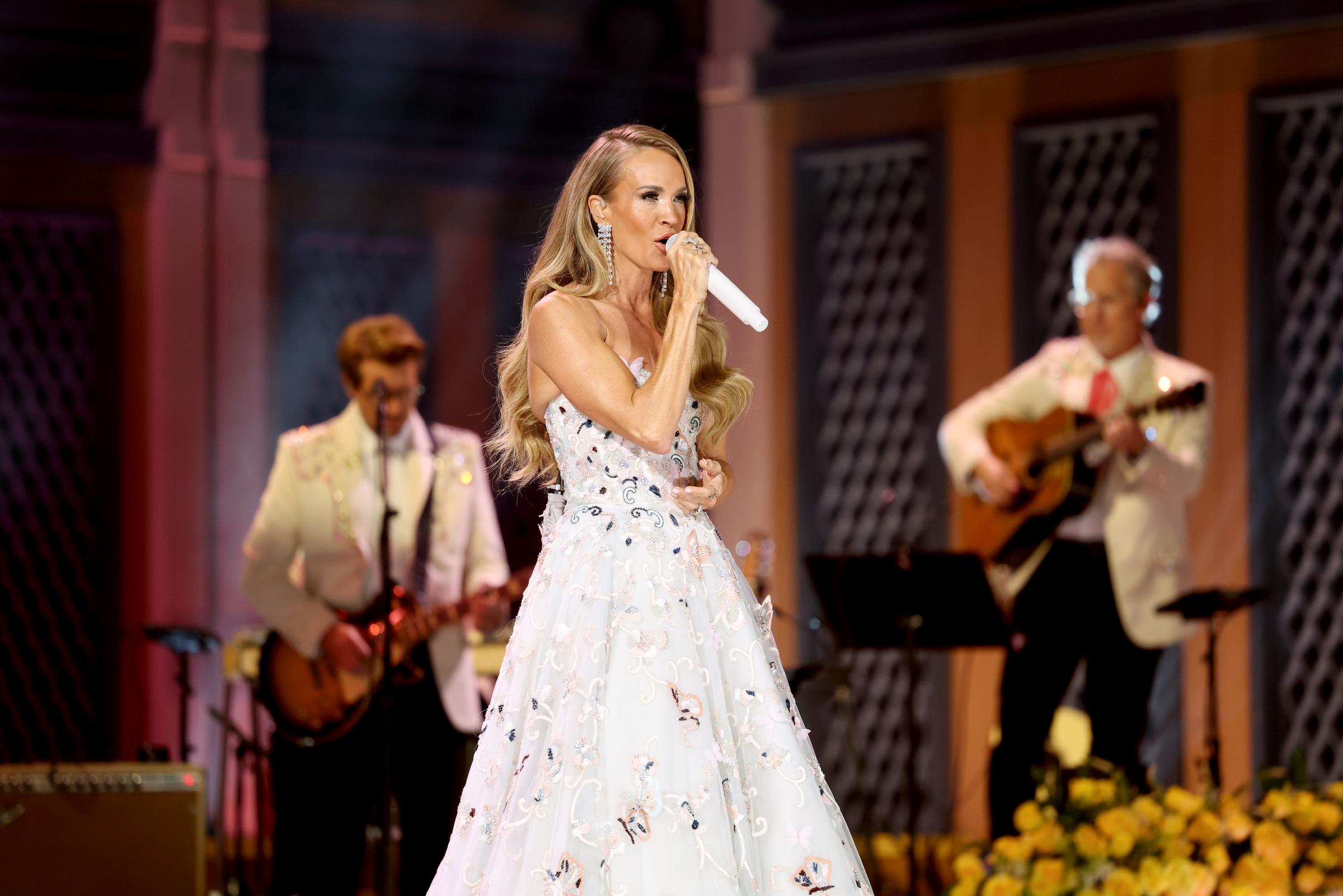 Carrie Underwood actúa junto a músicos vestidos con chaquetas blancas durante el homenaje a Dolly Parton, luciendo un vestido con bordados de mariposas como parte del homenaje de la noche a la leyenda del country. | Fuente: Getty Images.